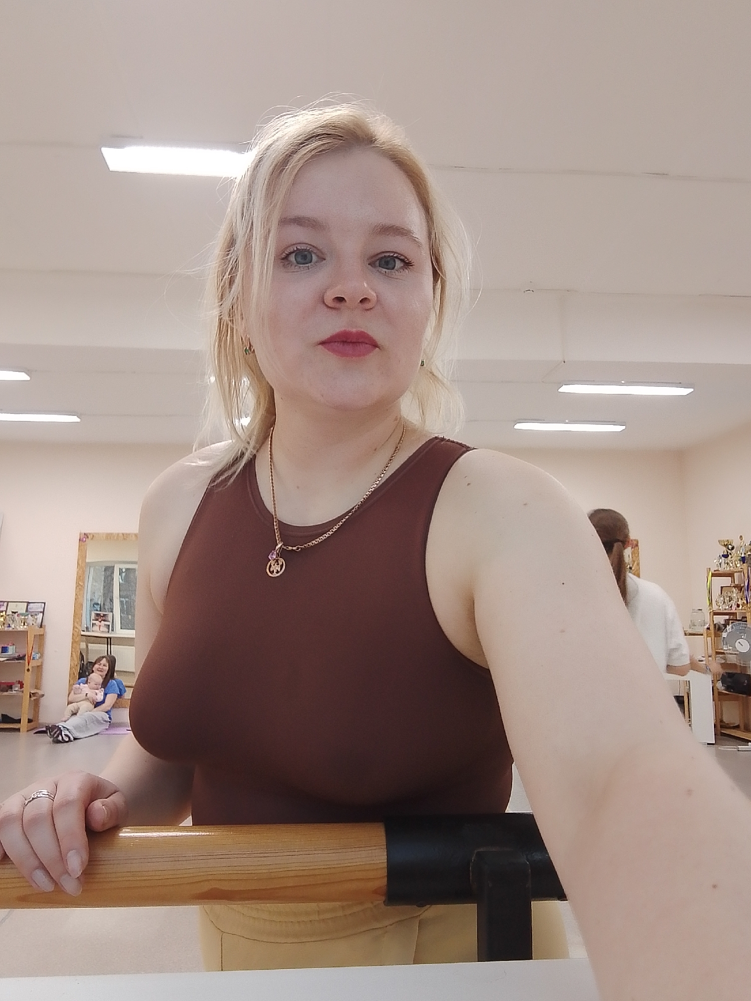 The image shows a fair-skinned woman with blonde hair, red lipstick, and blue eyes. She is wearing a brown tank top and a gold necklace with a pendant. A ring is visible on her left ring finger. She is standing at a ballet barre with her hand resting on it.
In the background, there are other people and ballet equipment, including a mirror, shelves, and trophies. The image was taken in Izhevsk, Russia.
