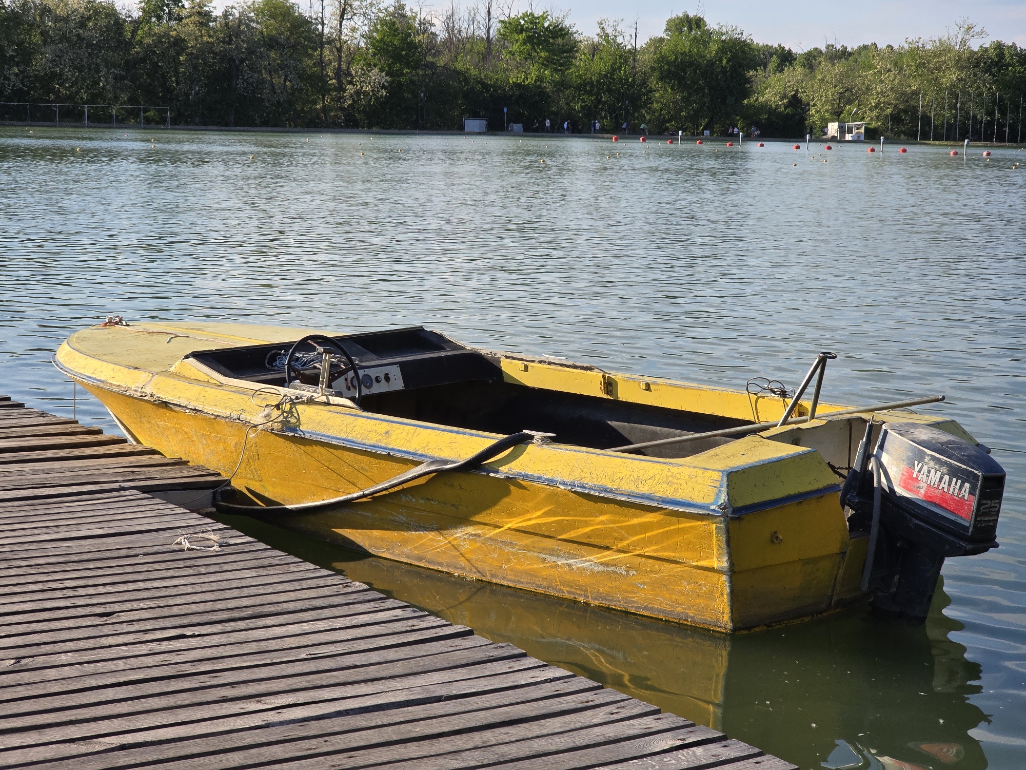 A yellow motorboat is docked alongside a wooden pier on a lake, near Unknown, Unknown. The boat is tied to the pier with a rope. The boat is equipped with a Yamaha 25 outboard motor. In the background, there is a row of trees, buoys, and some small structures on the opposite side of the lake. People can be seen in the distance near the structures. The water is slightly rippled.