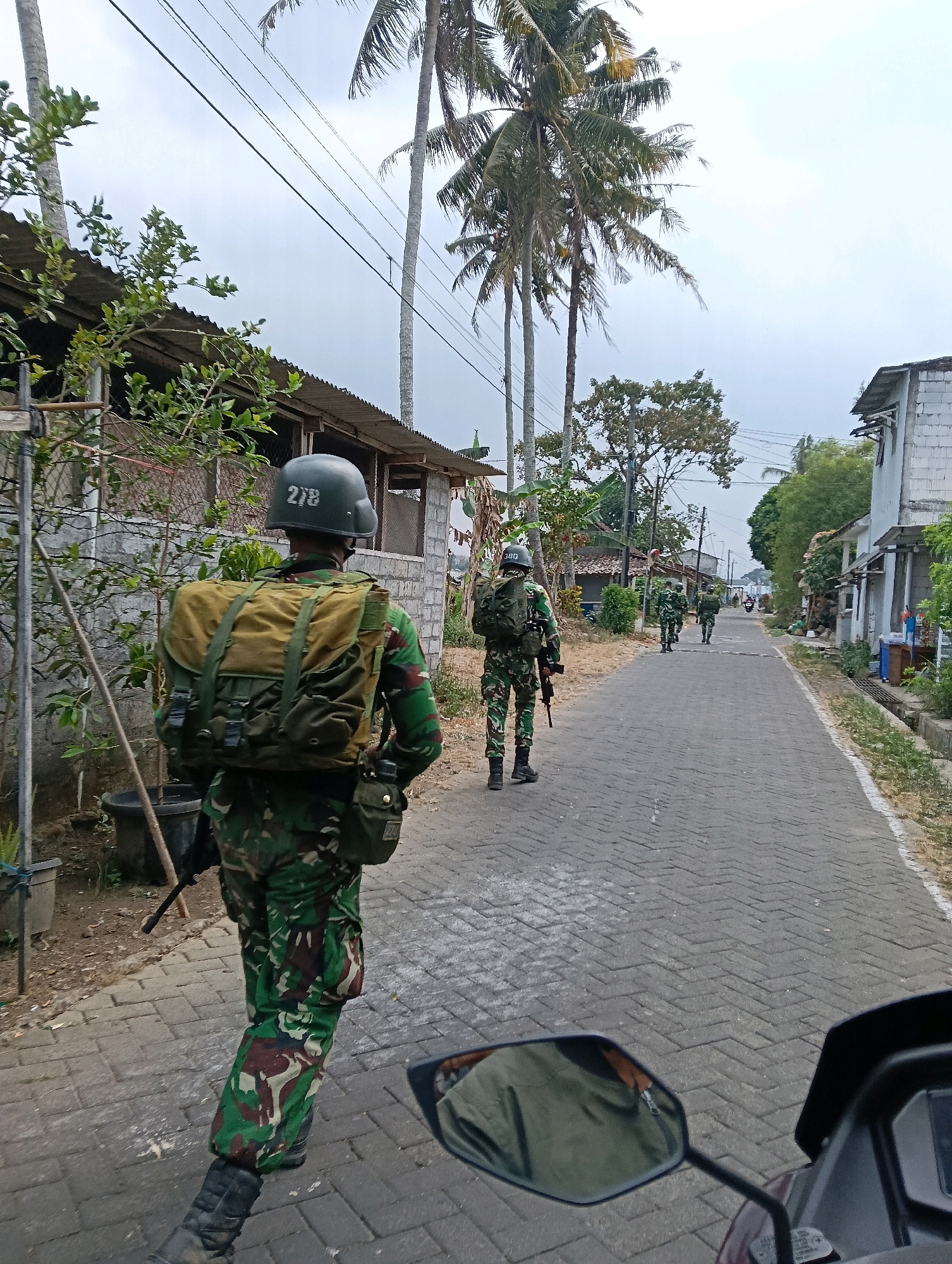 The image shows a group of soldiers in camouflage uniforms walking down a paved road in what appears to be a village or suburban area in Banyurojo, Indonesia. The road is lined with simple buildings and palm trees. The soldiers are equipped with backpacks and rifles. The perspective suggests the photo is taken from a vehicle, possibly a motorcycle, as a rearview mirror is visible in the foreground. The sky is overcast. One soldier in the foreground is facing away from the camera and has the number "278" on his helmet.