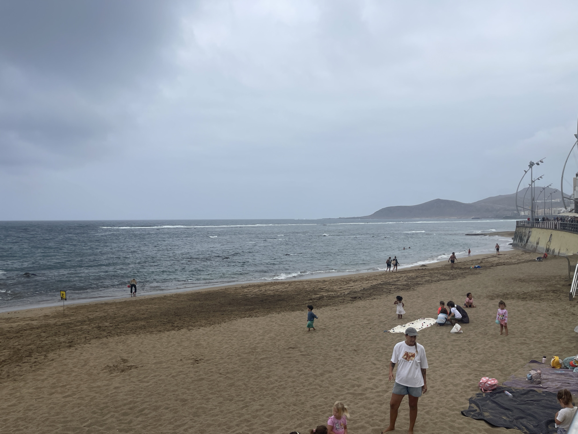This is a medium-shot, eye-level, outdoor, daytime, slightly overcast photograph of a sandy beach. The beach is a light brown color with some darker patches where the sand is wet. Gentle waves are breaking on the shore. The water is a dark blue color and there are some whitecaps. In the background, there is a faint outline of hills and mountains. Along the right side of the image, there is a promenade with streetlights and a wall with a yellow railing.
There are several people scattered across the beach. Near the water, a person in red shorts and a dark shirt is walking with a red bucket. Further down the beach, a small group of people are sitting on a white blanket, and other individuals are walking or standing. Closer to the foreground, a person wearing a white t-shirt and light-colored shorts is standing facing left, and a young child in a pink swimsuit is standing to the right of them. Another young child in a blue shirt and green shorts is running in the middle ground. To the far left, a person is standing near the water. There are also several bags and blankets scattered on the sand. The sky is a pale gray with some lighter patches, indicating overcast conditions.