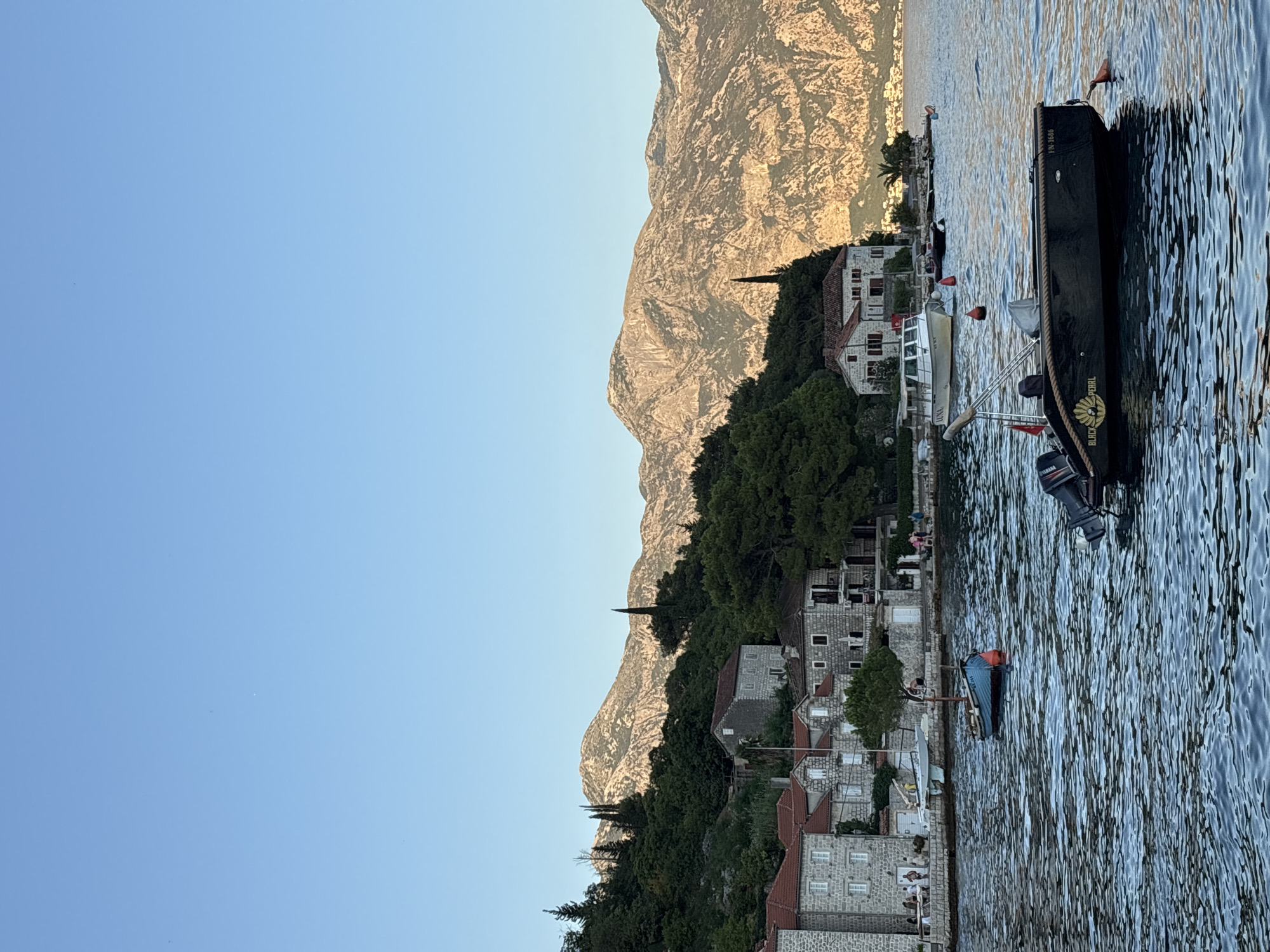The image captures a tranquil scene in Perast, Montenegro, a coastal town known for its historic architecture and stunning natural surroundings. The foreground is dominated by the calm, blue waters of the bay, with several boats gently bobbing on the surface. A prominent black motorboat, labeled "BLACK PEARL" and bearing the registration "FN-1686", is positioned in the center right, facing left. To its left, a smaller, blue rowboat with a red buoy is anchored.
In the mid-ground, the picturesque shoreline of Perast unfolds. Traditional stone buildings with red-tiled roofs line the waterfront, suggesting a historical settlement. Some of these buildings feature balconies and windows, hinting at residential or commercial use. Several people are visible along the promenade, some standing and others walking, enjoying the coastal ambiance. A white passenger boat, labeled "LUNA", is docked near the shore. Red buoys are scattered throughout the water, marking navigation points or moorings.
The background is defined by a lush, green hillside densely covered with trees, which rises to meet a majestic mountain range. The mountains, bathed in the warm light of what appears to be late afternoon or early morning sun, exhibit rugged textures and rocky outcrops, creating a dramatic contrast with the serene seascape and the charming town. The sky above is a clear, pale blue, devoid of clouds, and stretches to the horizon. The overall impression is one of peaceful coexistence between natural beauty and human habitation.