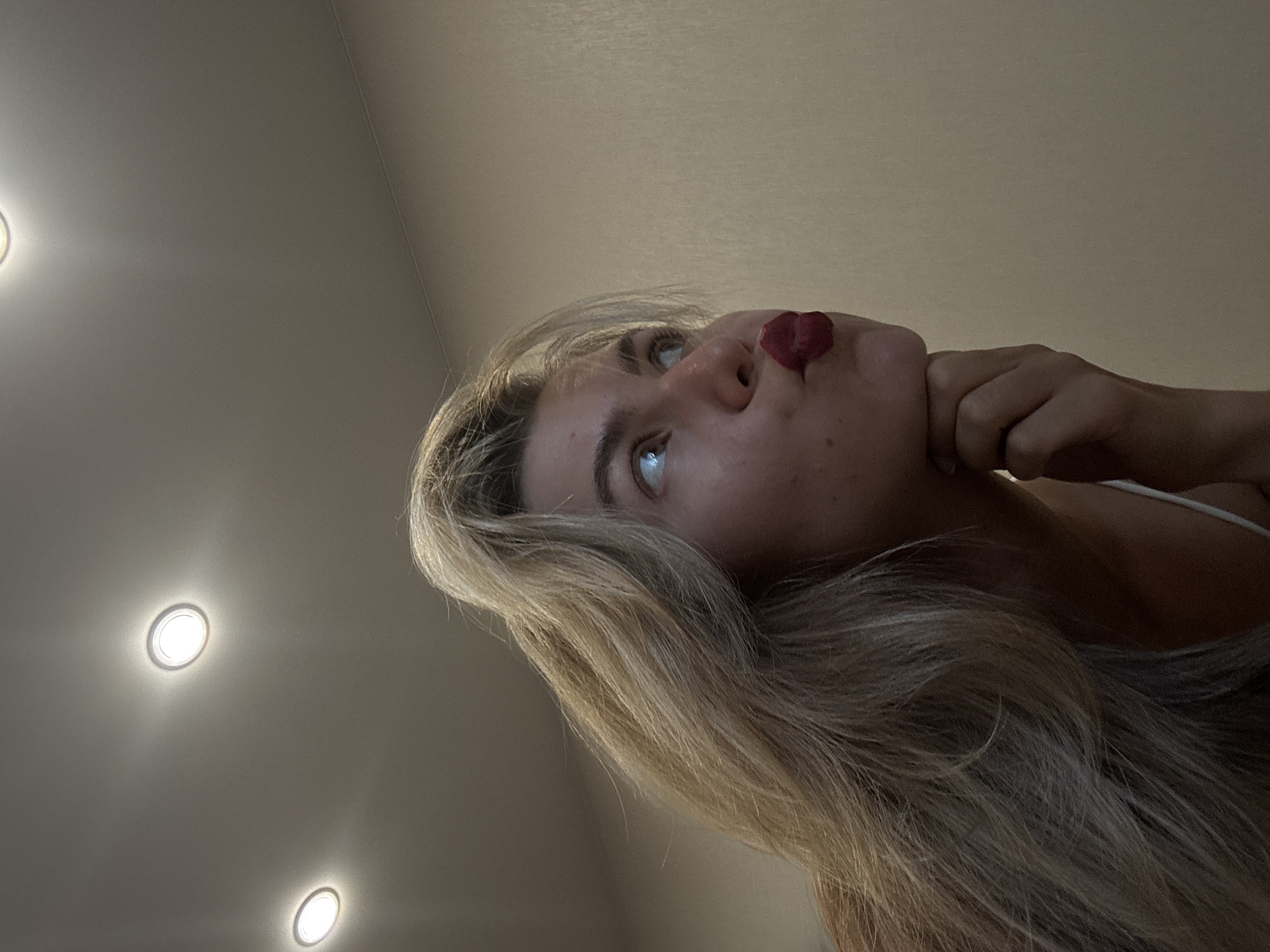 The image is a low-angle shot of a woman with blonde hair looking upwards. Her face is tilted back, and she has a pouting expression. Her right hand is touching her chin. Above her is a ceiling with recessed lighting, consisting of at least three circular lights. The location is in the city of Unknown, Unknown.