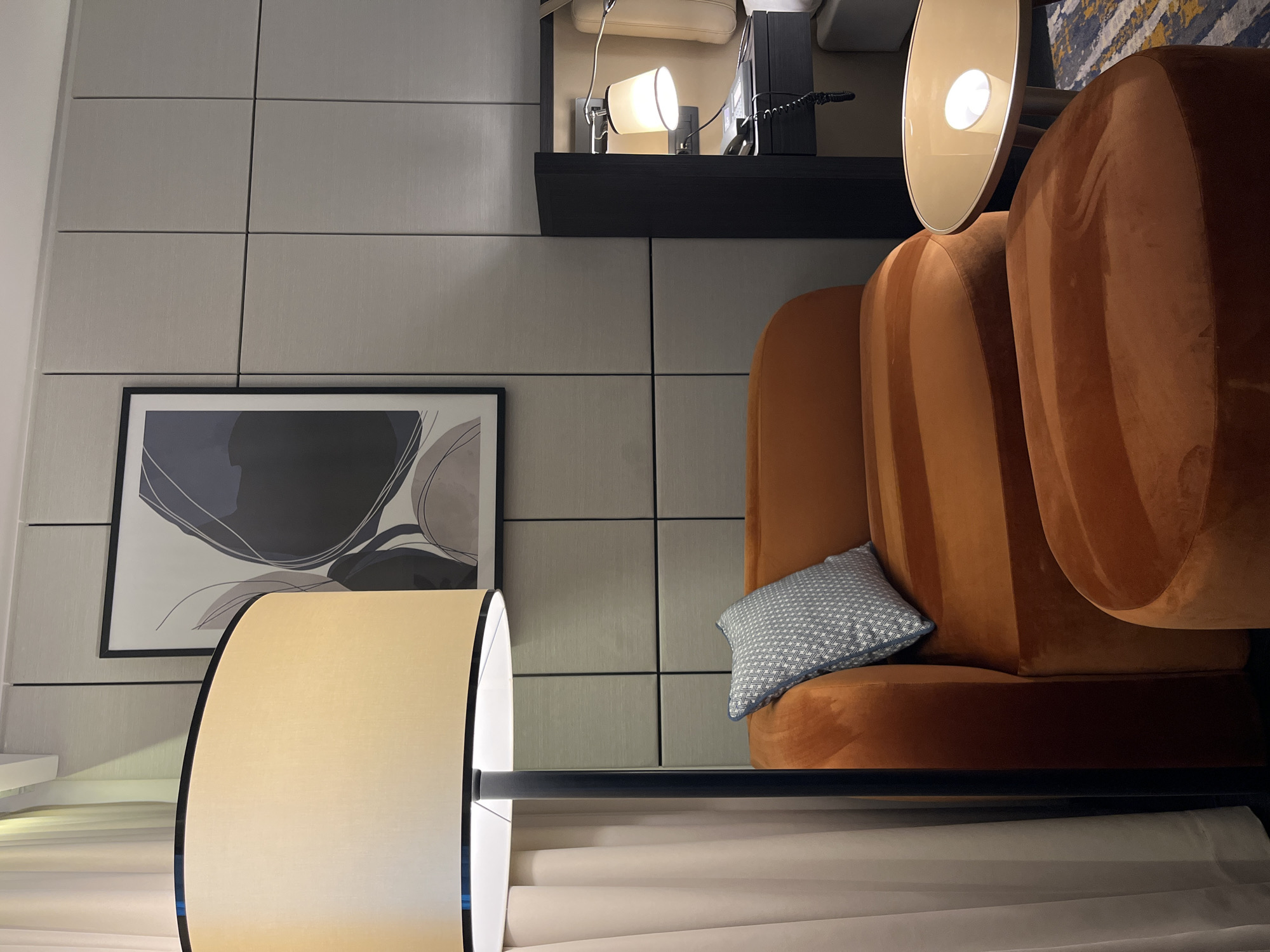 A room interior in Belgrade, Serbia is depicted. A burnt-orange velvet chair is prominently displayed with a patterned blue pillow resting on its seat. Behind the chair is a wall covered in grey, rectangular panels. A framed abstract artwork hangs on the wall to the left of the chair. A bedside lamp extends over the chair. A small black shelf is mounted on the wall above the chair, holding a telephone and a lamp. The room contains elements of a modern hotel or apartment.