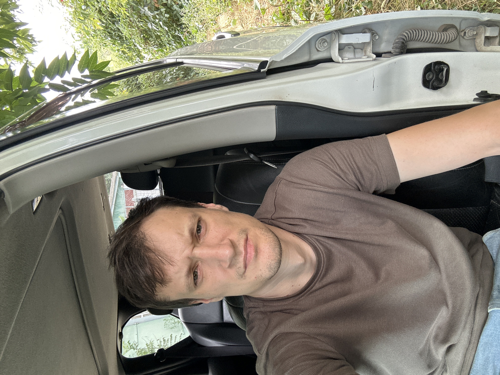 A man is sitting in the driver's seat of a white car, which is parked on a dirt road next to a dense area of green foliage. The car door is open, revealing the interior, which has black leather seats and a light gray headliner. The man is wearing a brown t-shirt and blue jeans. He has short, dark brown hair and is looking at the camera with a serious expression. The background shows trees, bushes, and a hint of a building. The location is identified as Makiv, Ukraine.