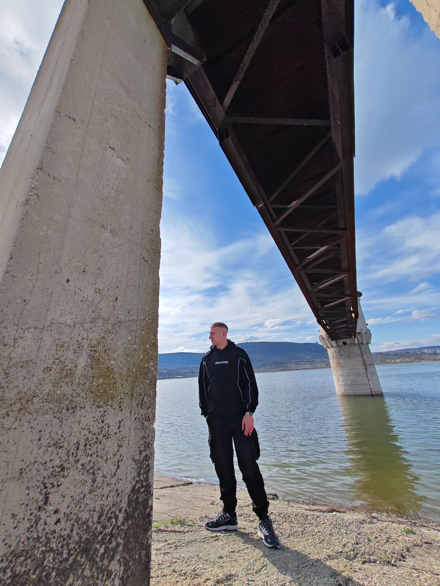 A man is standing under a bridge in Shumen, Bulgaria. He is wearing a black sweatshirt with white stripes, black pants, and sneakers. He is standing on a dirt and gravel shoreline of a body of water. The bridge is made of steel and concrete. The sky is blue with white clouds. A hill is visible in the background.