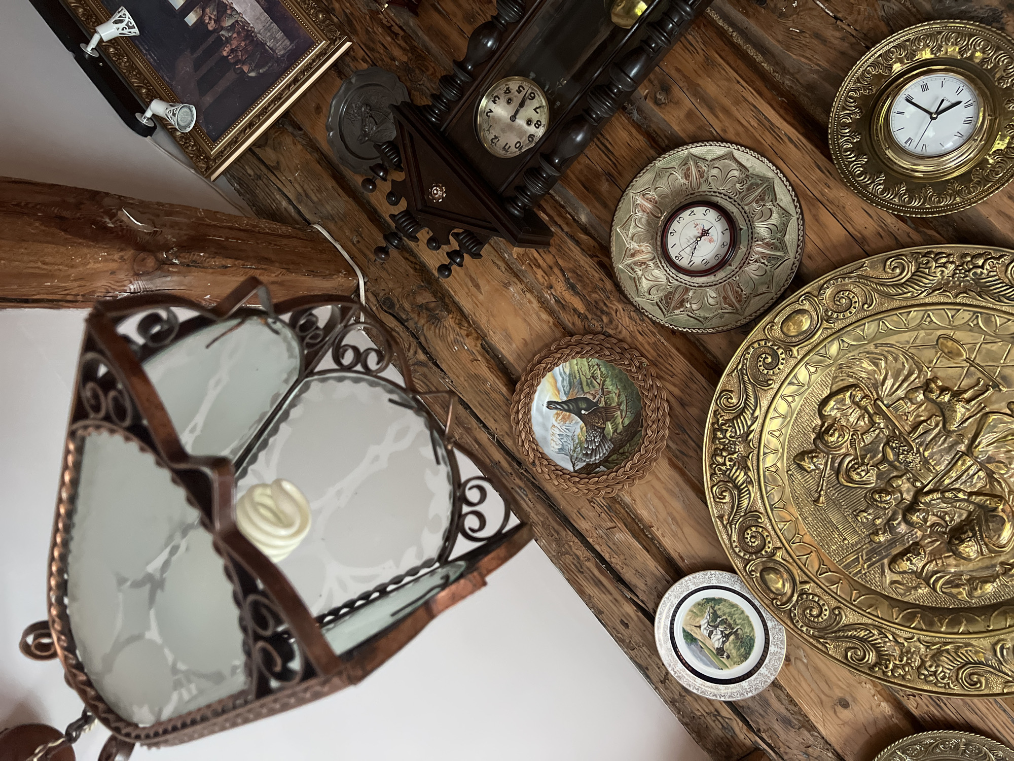 The image depicts an interior scene, likely within a rustic setting characterized by exposed wooden beams and a ceiling lamp. Several decorative objects are visible, including a large ornate metal plate with a relief scene, a wall clock with a dark wooden casing, and at least two smaller decorative plates with painted images of animals. A framed picture hangs on the wall. The overall impression is one of a collection of antique or decorative items displayed in a warmly lit, traditional environment. There is no indication of Norviliškės, Lithuania, within the image or its metadata.