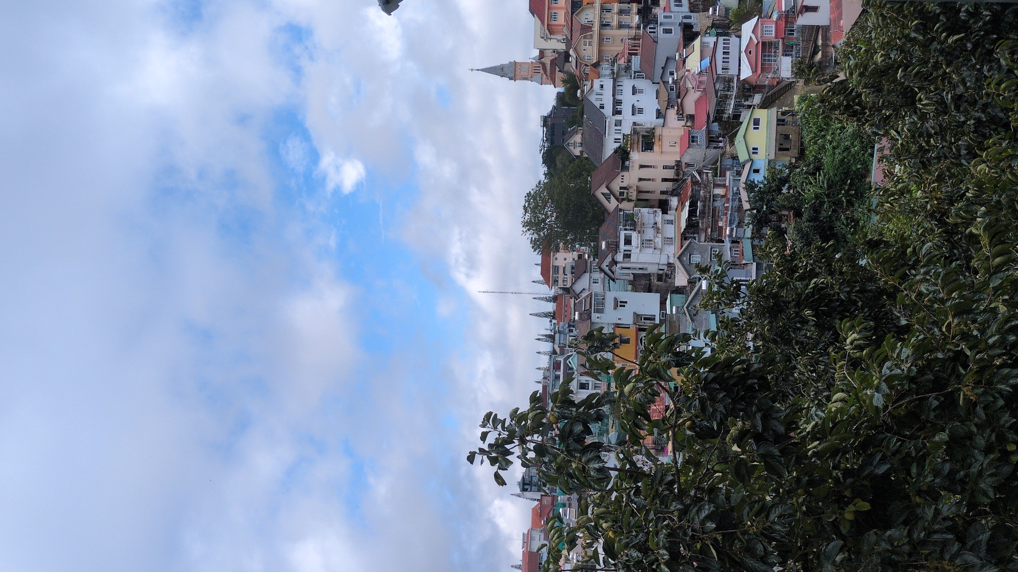 A wide-angle, eye-level shot captures a dense residential area in Da Lat, Vietnam, nestled on a hillside. The foreground is dominated by the dark green foliage of trees, partially obscuring the view of the town. The buildings are varied in style and color, with many featuring tiled roofs in shades of red, brown, and gray, and walls in white, yellow, and pastel hues. Some buildings have balconies and multiple stories. A prominent church steeple, constructed of a light-colored stone, rises above the rooftops to the right of the frame, indicating the presence of religious architecture within the city. A tall, slender communication tower is visible in the background. The sky above is a mix of bright blue and scattered white clouds, suggesting fair weather with some atmospheric cover. The scene depicts a typical urban landscape of Da Lat, characterized by its hilly terrain and diverse architecture. There are no people visible in the frame.