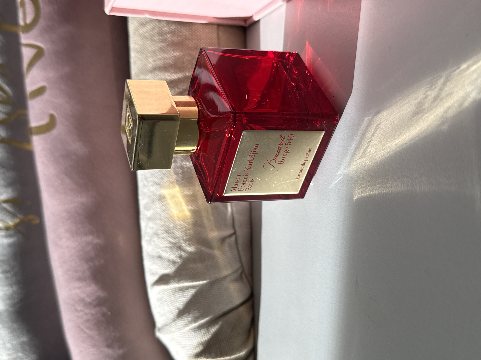 A red glass perfume bottle with a gold cap sits on a white surface. The bottle has a gold label that reads "Maison Francis Kurkdjian Paris Baccarat Rouge 540 Extrait de parfum". The background consists of folded pink and gray fabric. The scene is lit by sunlight casting shadows and reflections on the surface. The image does not contain any information about the city of Bad Sachsa, Germany.