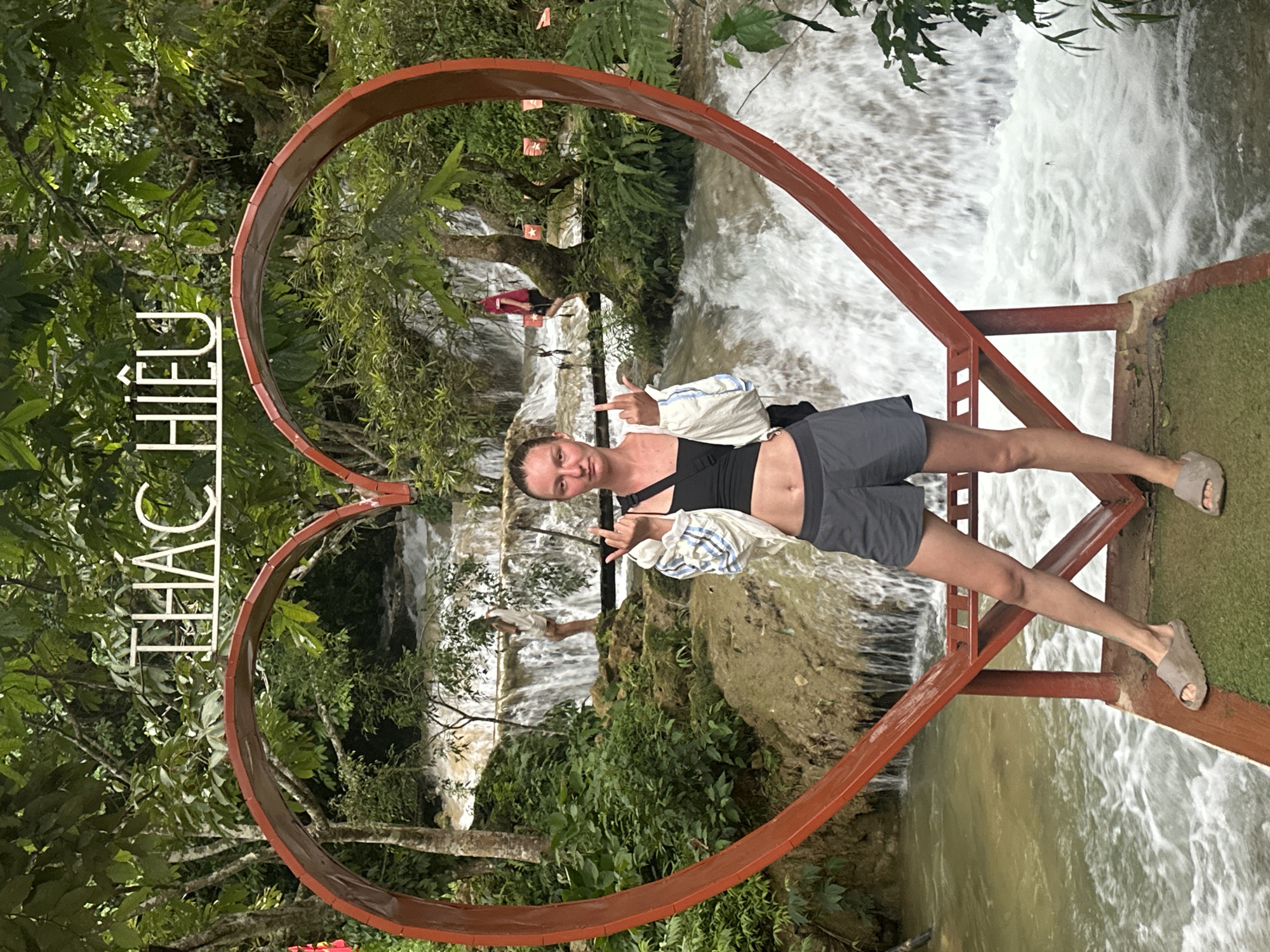 A young Caucasian woman stands in front of a red heart-shaped arch with a waterfall in the background. She is wearing a black crop top, grey shorts, and a light-colored jacket. Her hair is pulled back in a ponytail, and she is giving a middle finger to the camera. There is a sign with Vietnamese writing above the arch that reads "THÁC HIÊU". The waterfall is surrounded by lush green vegetation, and the water appears to be a light brown color. In the background, there are two other individuals, one standing on a rock formation near the waterfall and another partially visible further back. The scene is located in Cổ Lũng Commune, Vietnam.