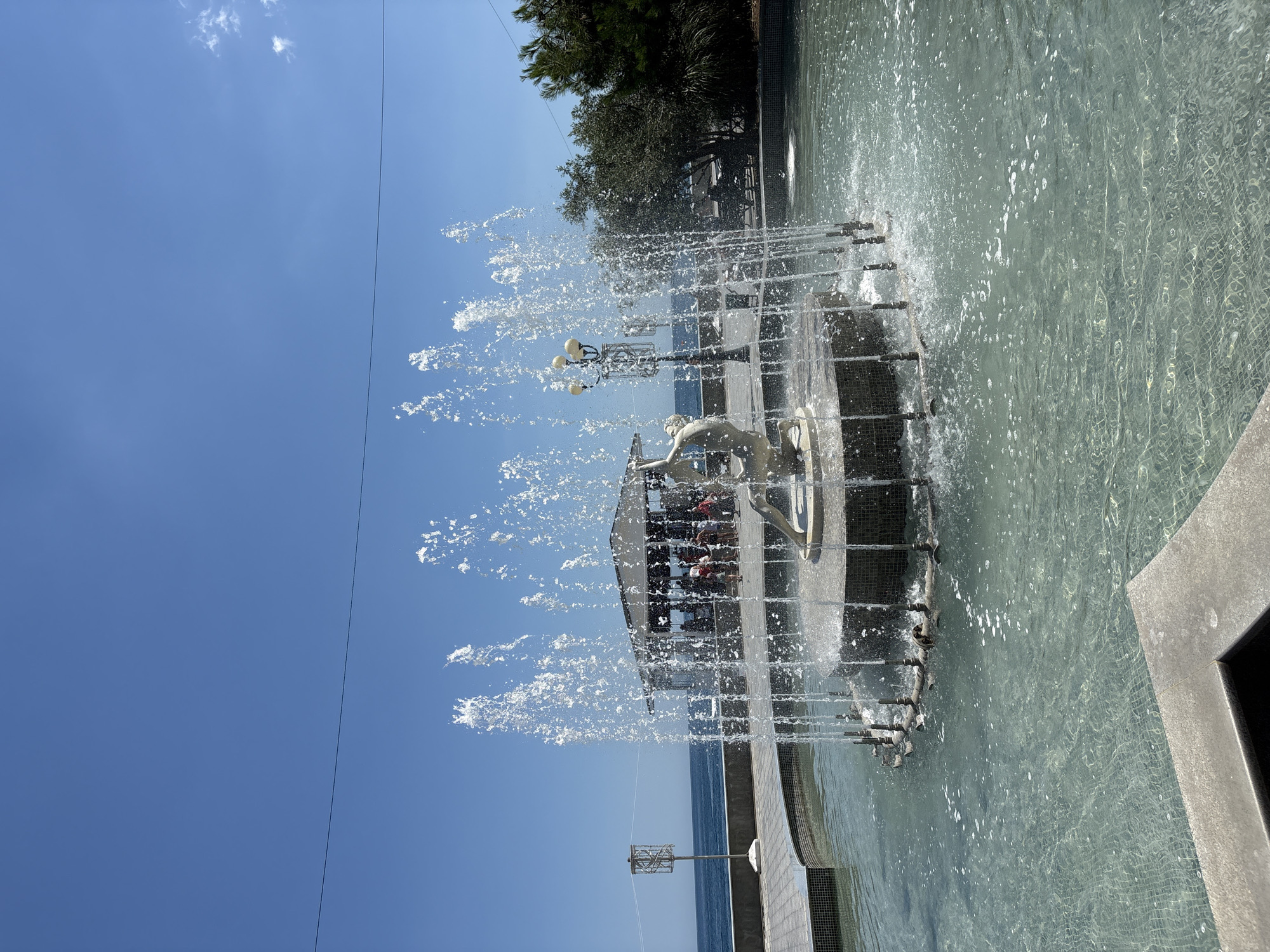 The image shows a fountain in a park setting with the Black Sea in the background. The fountain features a sculpture of a person in the center, surrounded by water jets. There are people in the background, some of whom appear to be dining at an outdoor cafe. The park also has trees and benches. The sky is clear and blue, indicating a sunny day. The location is identified as Alushta, Ukraine.