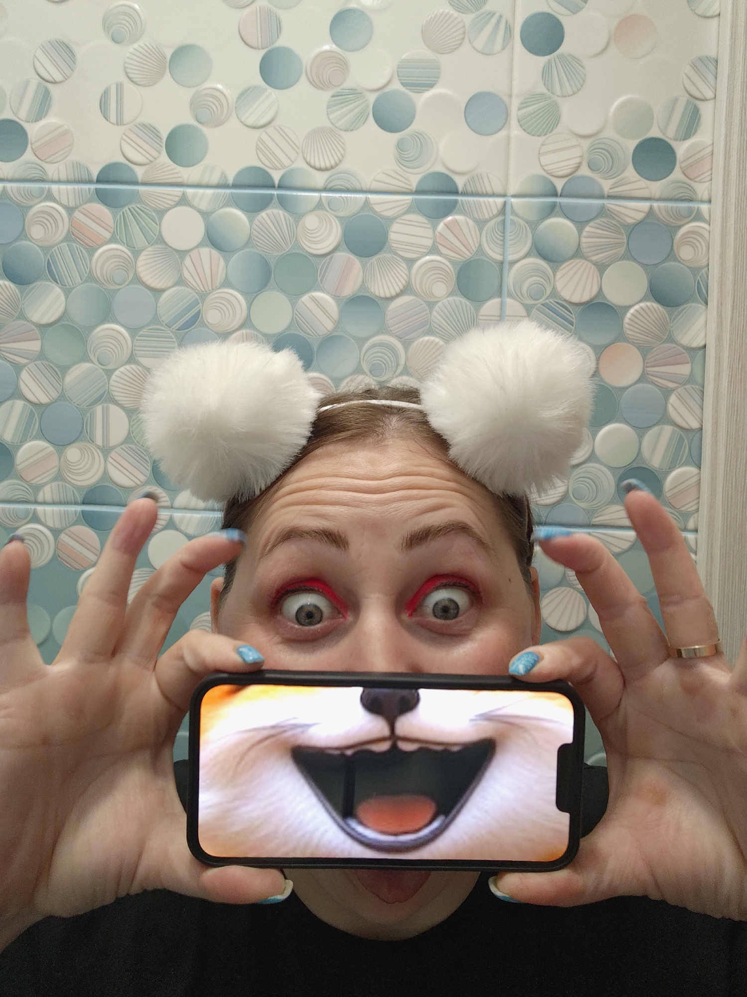 An indoor, eye-level shot in Markovskii, Russia, captures a person humorously interacting with a smartphone. The subject wears a headband adorned with two large, fluffy white pom-poms, positioned above her head. Her eyes are wide open, emphasized by striking bright red eyeliner or eyeshadow, giving her a look of exaggerated surprise. Her hands, featuring light blue nail polish and a simple gold ring on her right ring finger, hold a black-cased smartphone horizontally, directly in front of her mouth and chin, obscuring them.
The smartphone screen displays a vibrant, close-up image or video of a light-furred animal, resembling a fox or dog, with its mouth wide open, revealing teeth and a tongue, suggesting a joyful or playful expression. The background consists of a tiled wall featuring a mosaic pattern of numerous circular tiles in varying shades of light blue, white, and soft pink. Many of these tiles have subtle striped or swirled textures, creating a decorative and bright setting, likely a bathroom or kitchen. The overall impression is one of lighthearted fun and creativity. There is no visible text in the image.
