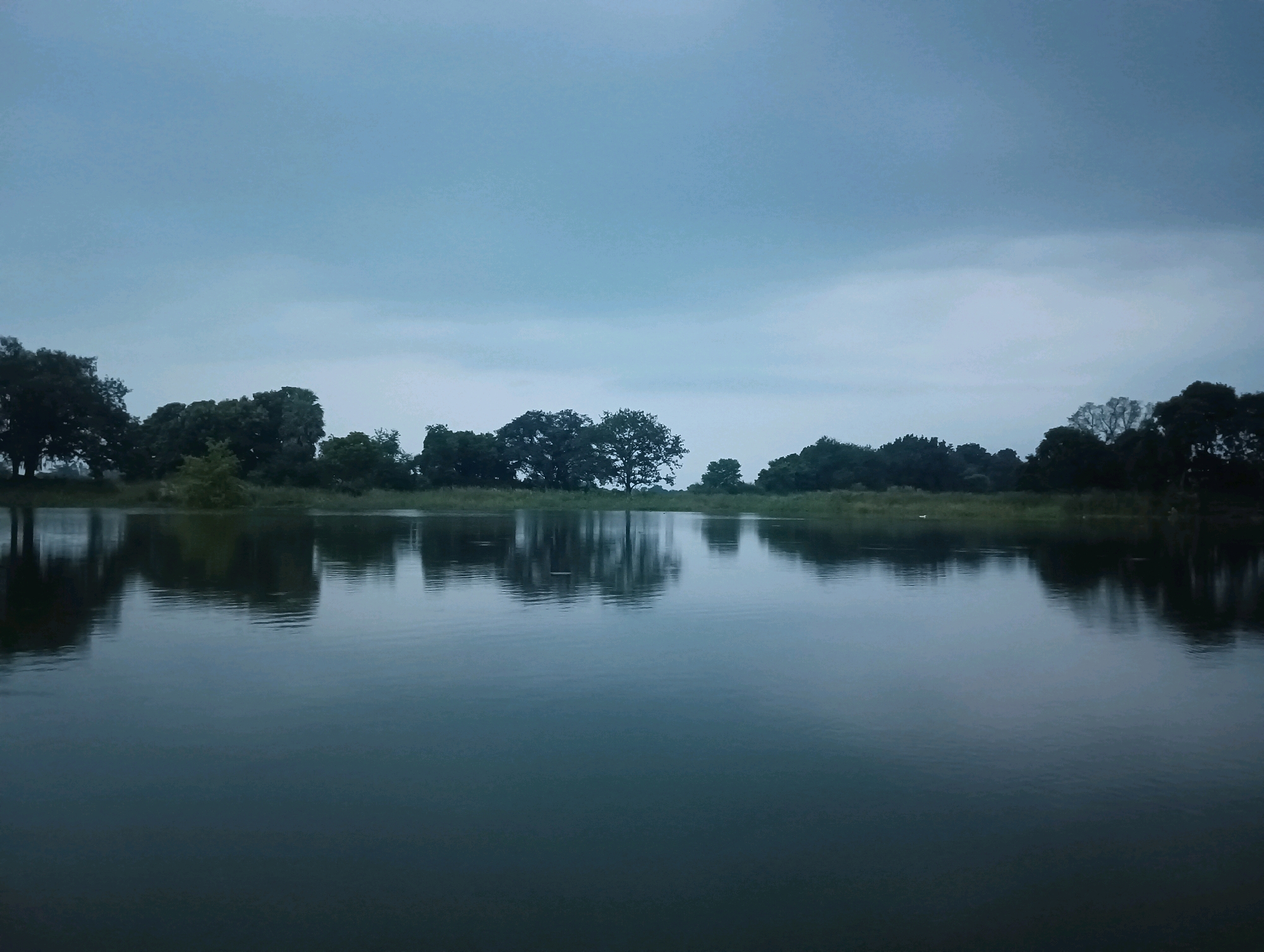 The image depicts a serene natural landscape with a body of water in the foreground, reflecting the cloudy sky and a line of trees along the horizon. The scene appears to be a natural setting, likely a lake or river. The sky is overcast with a soft, diffused light, contributing to the tranquil atmosphere. The water is still, creating clear reflections of the trees and sky above. The trees on the far bank are lush and green, forming a dense border against the pale sky. The overall impression is one of peace and natural beauty, possibly captured during dawn or dusk due to the muted light. There are no people or man-made structures visible, emphasizing the untouched quality of the environment. The location is identified as the city of Tilkar, India.