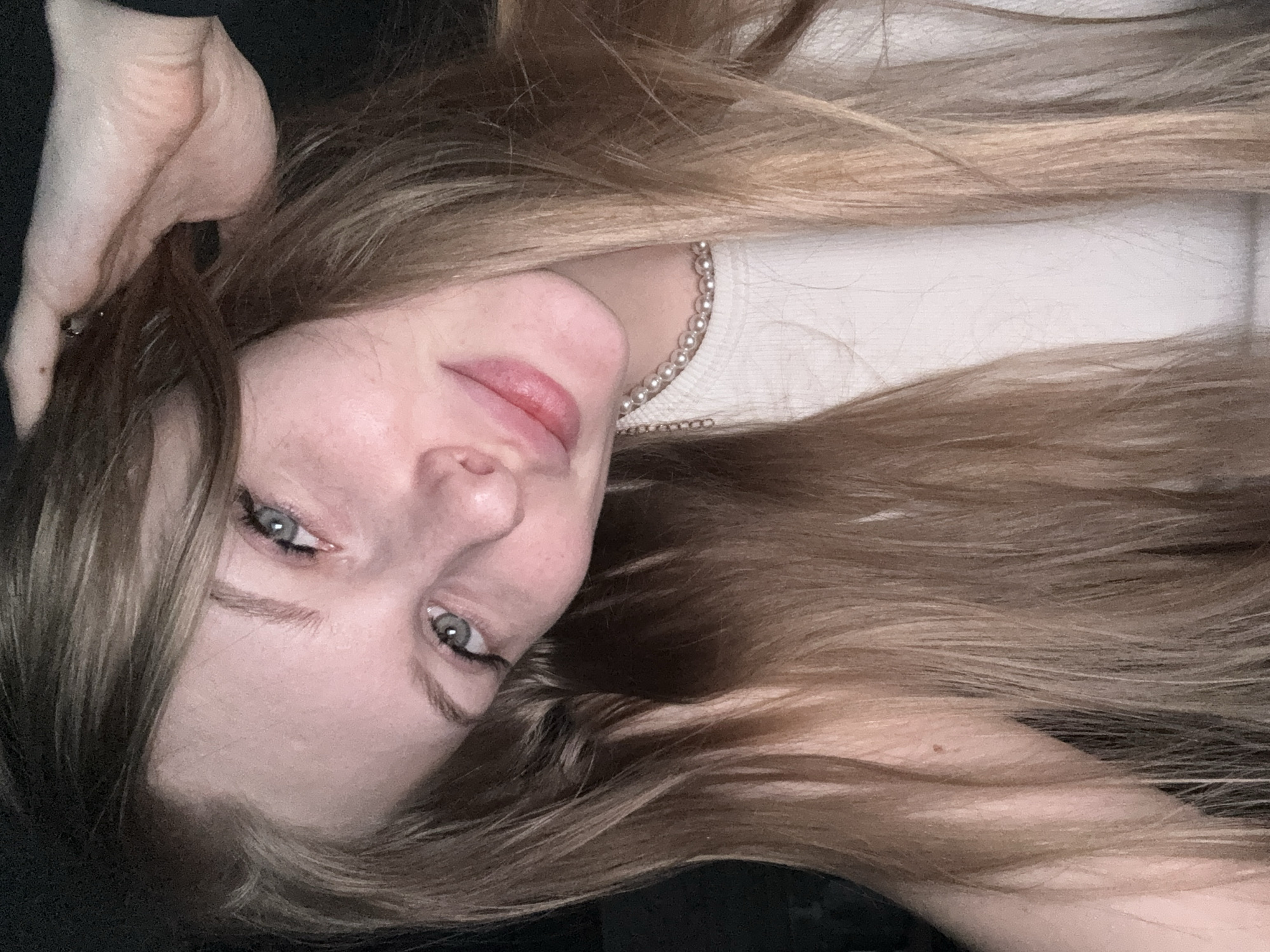A close-up, tilted portrait depicts a young woman with long, flowing light brown hair, accented with lighter highlights, framing her face and spread across a dark background. Her head is angled to her left, and she looks directly at the camera with light-colored eyes, possibly blue or grey, highlighted by black eyeliner. A subtle, closed-mouth smile is visible on her lightly colored lips. Around her neck, she wears a pearl necklace that rests above a white, ribbed top.
A notable detail is a light-colored, fleshy, ear-shaped object, resembling a pig ear prop or costume accessory, visible on the upper left side of the frame, seemingly positioned near her left ear and emerging from her hair. The lighting is soft and even, highlighting her features against the predominantly dark surroundings. The setting appears to be indoors, likely in Perm, Russia, given the provided context. The woman's expression is calm and engaging. No text is visible in the image.