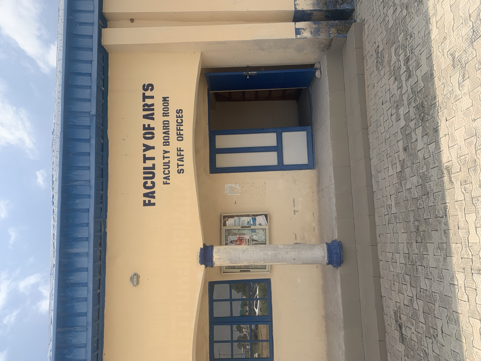 The image shows the exterior of the Faculty of Arts building at a university in Jalingo, Nigeria. The building is painted a pale yellow and has a blue corrugated metal roof. Large black stencil letters on the facade read "FACULTY OF ARTS", "FACULTY BOARD ROOM", and "STAFF OFFICES". To the left is a blue-framed window with multiple panes, reflecting cars and buildings outside. To the right of the window is a noticeboard with posters and flyers. A blue and white striped pillar stands in front of the building. A double blue door, one leaf open, leads into the building. The ground in front is paved with interlocking grey bricks. The sky is partly cloudy.