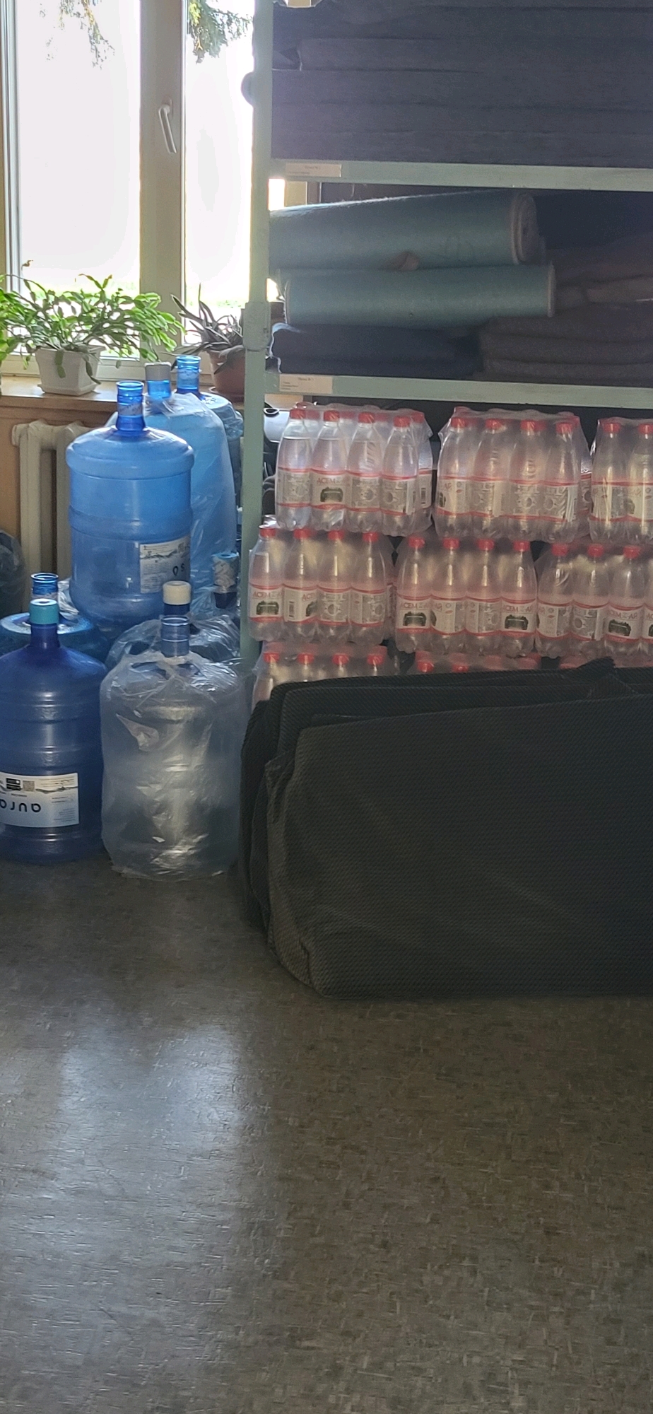 This image shows an indoor storage area. In the foreground, there are several large blue plastic water bottles, some of which are capped with blue and white plastic stoppers. One of these bottles, on the left, has a label with text that appears to read "AURO". Another large water bottle to its right is partially covered in clear plastic. Behind these are more blue water bottles, some with their plastic wrappings still attached. Further to the right, a stack of plastic-wrapped bottles of clear liquid, likely water, are arranged in rows. These bottles have red caps and labels with text that appears to read "ACEM". In the middle ground, a dark green fabric item, possibly a bag or a folded piece of material, is visible. In the background, a metal shelf unit is filled with rolls of fabric, appearing to be in shades of dark blue and light blue-green. A window is visible on the far left, with sunlight streaming through it, illuminating some of the scene and casting shadows. A potted plant with green leaves sits on the windowsill. The floor appears to be linoleum or a similar hard surface with a textured pattern. The scene appears to be a storage room or possibly a room in a dormitory or an educational institution, given the presence of water bottles and rolls of fabric, which could be used for upholstery or crafts. The image is taken in the city of Oskemen, Kazakhstan.