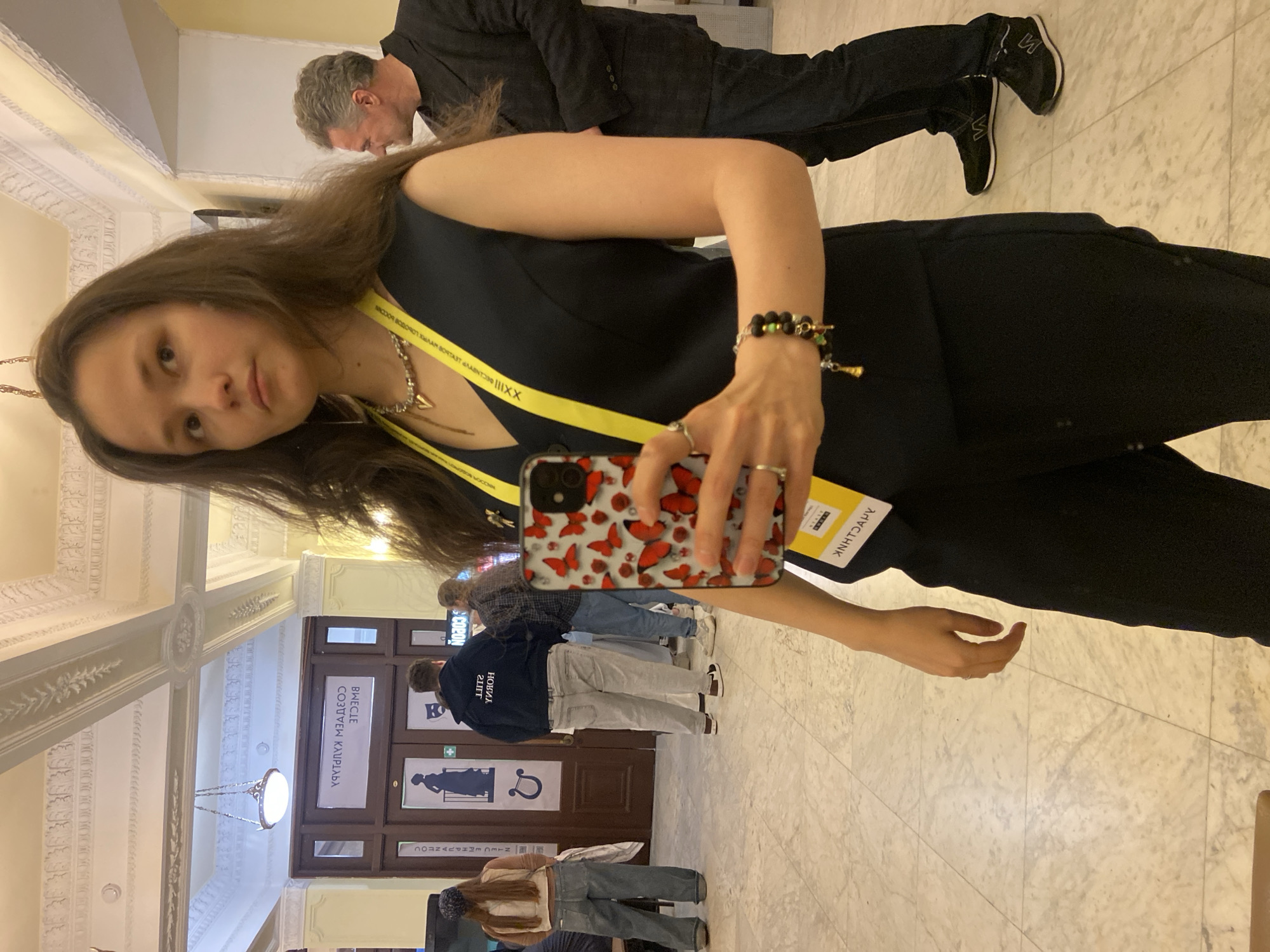 An indoor, slightly rotated image captures a young woman in the foreground, taking a selfie or being photographed. She has long brown hair and is wearing a black sleeveless top or dress. A yellow lanyard hangs around her neck, displaying text. She holds a smartphone with a distinctive red butterfly case in her right hand, adorned with rings and bracelets on her wrist. In the upper background, an older man with grey hair, dressed in a dark suit and black sneakers, walks away.
The setting is a grand hall or corridor, characterized by a light-colored, patterned marble floor and an ornate, decorative ceiling with chandeliers. Large wooden double doors are visible in the background, featuring various signs and posters. Other people, partially obscured, can be seen near these doors.
The yellow lanyard on the main subject contains Cyrillic text, including "XXIII МЕЖДУНАРОДНЫЙ КИНОФОРУМ ЗОЛОТОЙ ВИТЯЗЬ" which translates to "XXIII International Film Forum Golden Knight," indicating she is attending this event. Additional visible text on the lanyard and a yellow tag includes "XXIII," "АННТОАРУ" (ANHTOARU). Signs on the doors bear Cyrillic text such as "ВМЕСТЕ СОЗДАЕМ БУДУЩЕЕ" (Together We Create the Future), "ОТКРЫТЫЙ МИР" (Open World), and "ЗОЛОТОЙ ВИТЯЗЬ" (Golden Knight), along with symbols like a person's silhouette and a horseshoe. These details align with the context of Sergiyev Posad, Russia, which hosts the Golden Knight International Film Forum. The overall impression is that of an attendee at a cultural or public event.