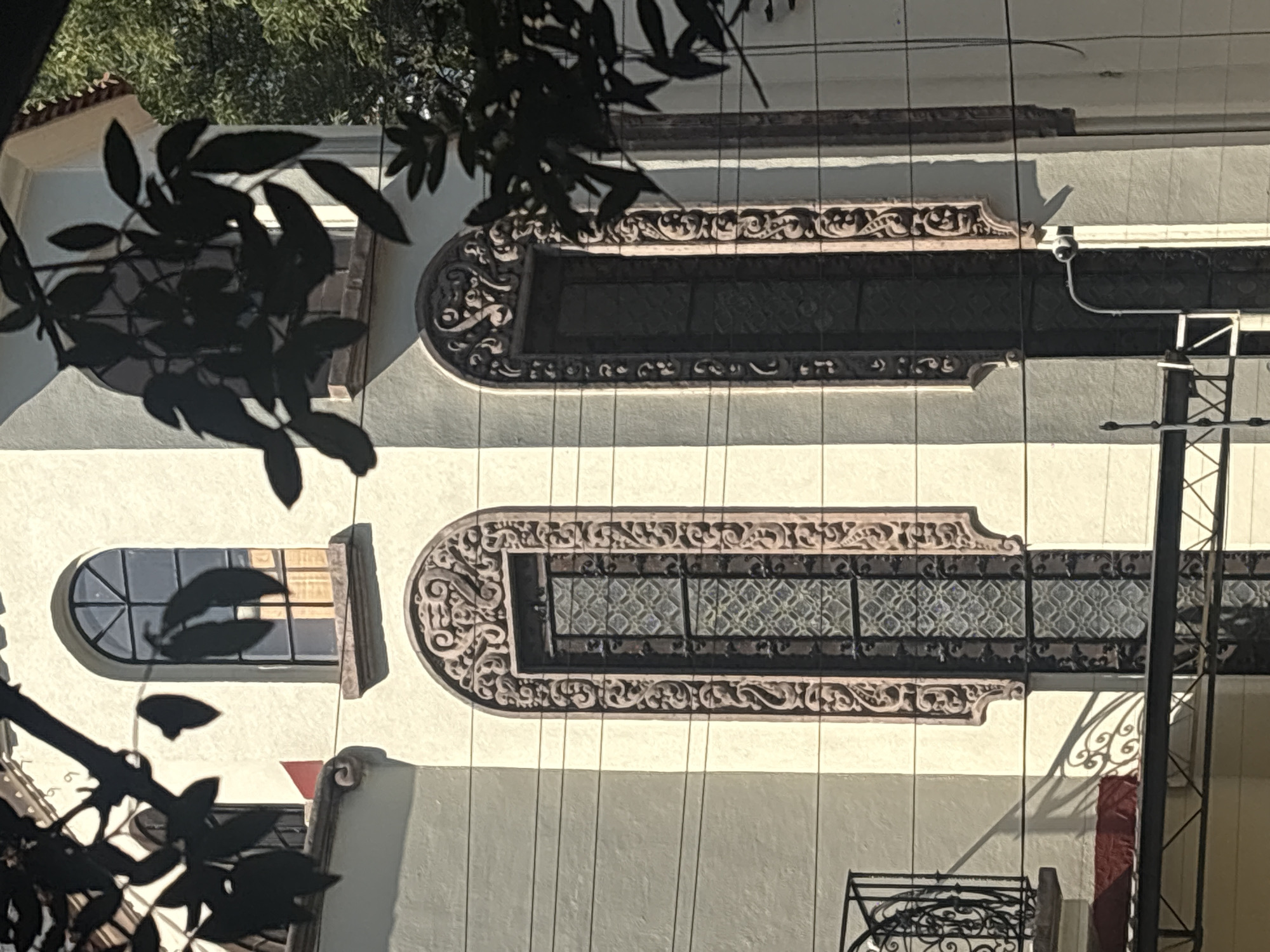 A vertical, eye-level shot shows a building facade in Mexico City, Mexico. The building is light beige, possibly stucco. Architectural details include two long, horizontal windows framed with elaborate, scrolled carvings above and below. One window to the left is arched at the top. A security camera is mounted on the building between the two windows. An electrical pole and lines are visible against the building. Parts of a wrought iron balcony are at the bottom right. A dark plant with leaves obscures part of the image. The light suggests the shot was taken during daylight.