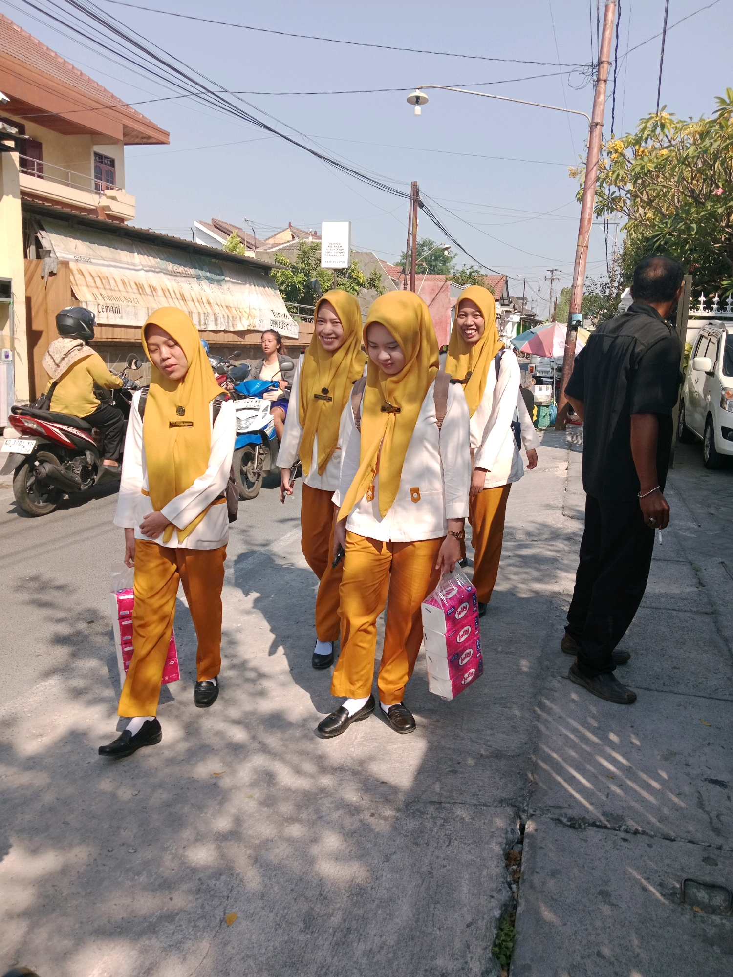 In Cemani, Indonesia, four young women in yellow hijabs and white tops with orange accents walk on a paved street under a clear sky. Two of them carry stacks of pink and white tissue boxes. A man in a black shirt and black pants walks on the sidewalk to their right. Several motorcycles are parked on the side of the road, one of which has a rider wearing a yellow headscarf and patterned scarf. Buildings with tiled roofs line the street, and overhead power lines crisscross the sky.