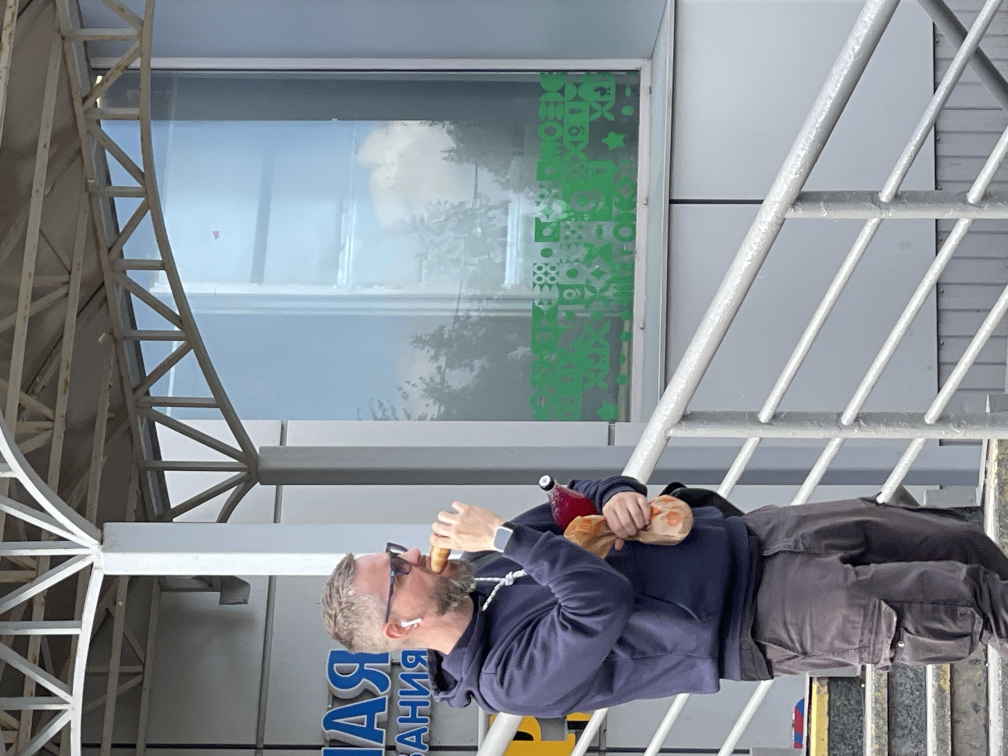 The image shows a man eating a sandwich and holding a drink while standing on a staircase. He is wearing a dark blue hooded sweatshirt, dark gray cargo pants, and sunglasses. He has a beard and is wearing white earbuds. The drink appears to be in a glass bottle with a red liquid. He is holding a brown paper bag in his left hand, which also contains a sandwich.
The setting appears to be outdoors, near a building with a large window. The window has a green geometric pattern on it. There is a metal structure with diagonal bars behind the man, possibly part of a staircase railing or awning. The sky is visible through the window, suggesting it is daytime. The overall lighting indicates it is likely a clear or partly cloudy day.
The man's action of eating suggests a casual moment, possibly a lunch break or snack. His body language is relaxed. The background details, such as the building and the staircase, suggest an urban environment. The visible text on a sign below the man reads "ЯСНАЯ" which is a Russian word meaning "clear" or "bright". The context provided that the location is Moscow, Russia, further grounds the scene.