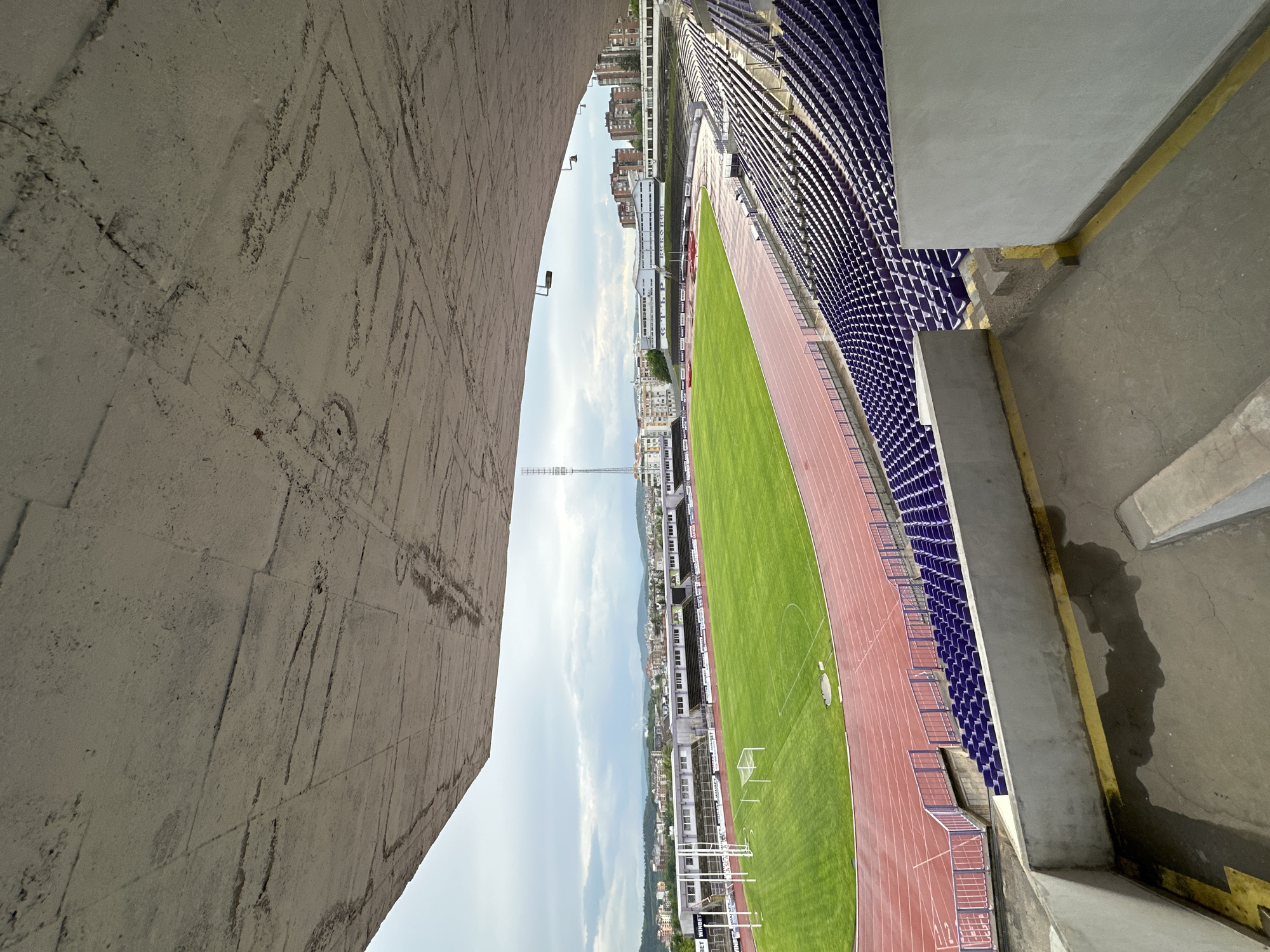 The image shows a partial view of the Ivaylo Stadium in Veliko Tarnovo, Bulgaria, from an elevated position. A large concrete arch is visible on the left side of the frame. In the center, a green soccer field is surrounded by a red running track. Rows of blue stadium seats are visible on the right. In the background, a cityscape is visible, with buildings and a tall communication tower. The sky is overcast.