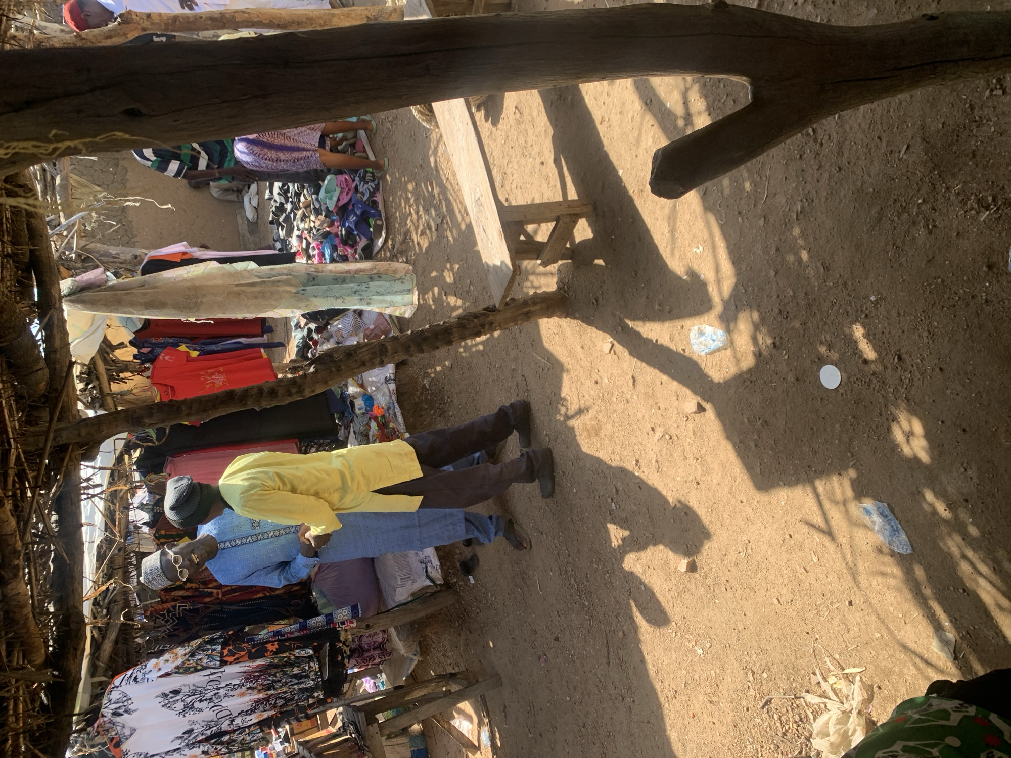 The image depicts an outdoor market scene in Dull A, Nigeria, bathed in the bright sunlight of what appears to be midday. The foreground is dominated by rough-hewn wooden structures, likely part of a stall or awning, casting long shadows across the dusty ground.
The central subjects are two men standing near a collection of colorful textiles and clothing. The man on the right is wearing a bright yellow robe and a dark beanie, with his head tilted slightly downwards. To his left, a man in a blue traditional outfit and a patterned cap is looking towards the camera, wearing sunglasses. His expression appears neutral or perhaps slightly amused.
Behind the men, various garments are displayed, including patterned dresses and other clothing items hanging on racks or folded. The ground is dry and sandy, with some scattered debris, including a few plastic wrappers and a small white object. In the background, more market stalls and goods are visible, suggesting a bustling commercial environment. The overall impression is of a vibrant, casual market day. There is no visible text in the image.