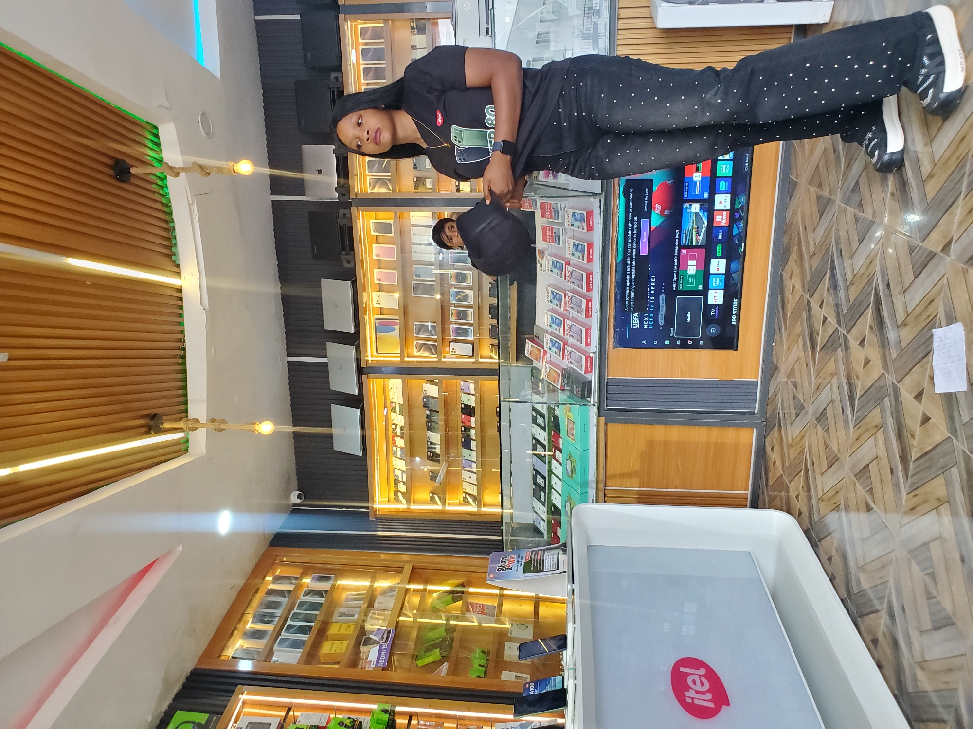 In an electronics store in Jos, Nigeria, a woman stands near a display of phones, holding a black bag. She is wearing a black t-shirt with a logo and black pants adorned with rhinestones, along with sneakers. Behind her, shelves are filled with mobile phones, and a man is partially visible in the background. The store features wood-paneled walls and ceiling with integrated lighting, as well as a large television screen displaying a media interface. The floor is tiled with a patterned design, and a white counter with a logo is in the foreground.
