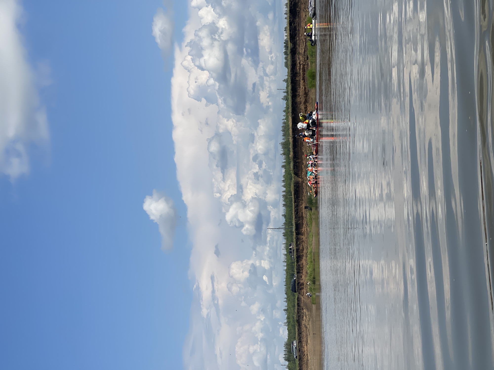 In a wide, calm river under a partly cloudy sky, a group of people are on a raft in the water. The raft is made of several inflatable kayaks and paddleboards tied together. Approximately ten people are visible on the raft, some standing and some sitting. They are wearing casual clothing, with some individuals wearing orange or yellow life vests. The riverbank on the far side is visible, with a brown, eroded cliff face topped by a line of green trees. A few vehicles are parked on top of the cliff. To the right of the raft, two smaller inflatable boats are also on the water. The scene appears to be a recreational outing on a river, possibly near Nezhinka, Russia, given the typical landscape and activity.