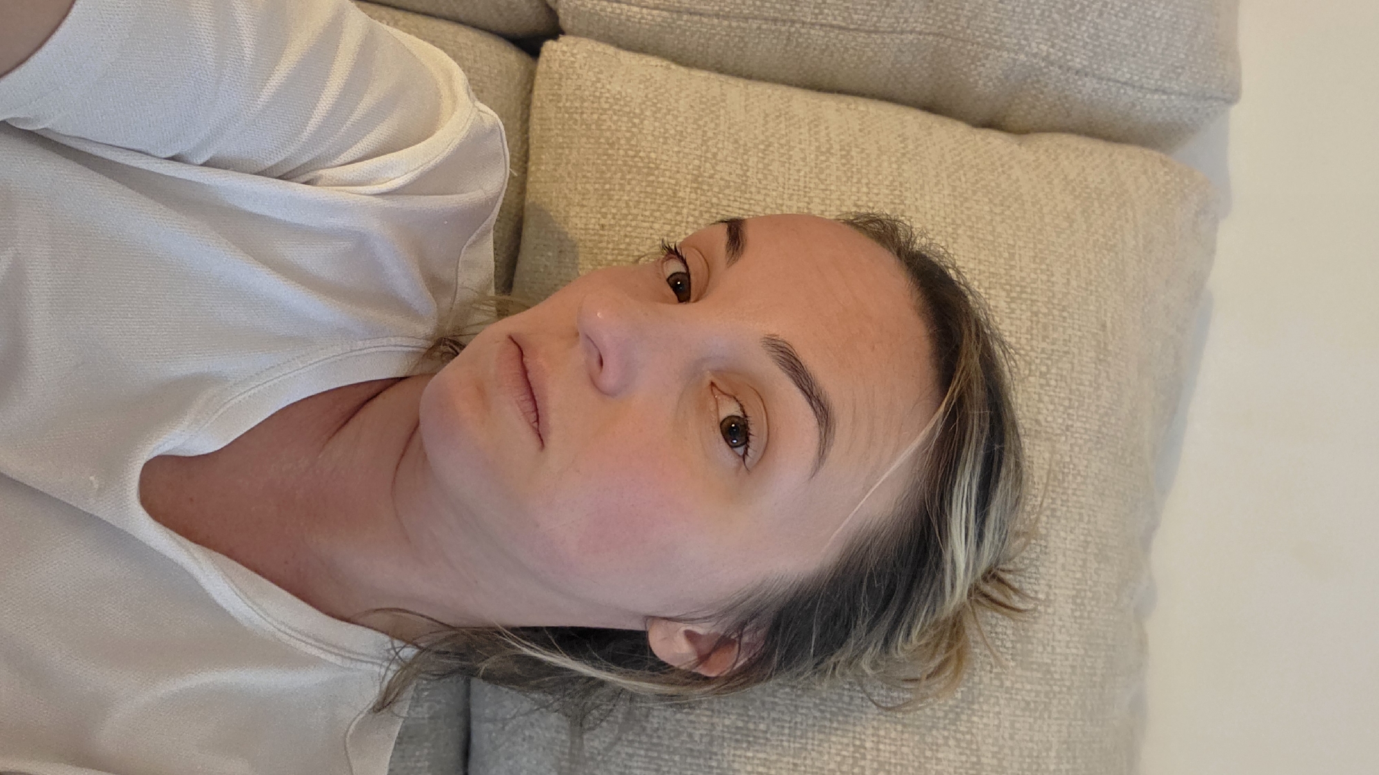 A young woman with blonde hair pulled back in a bun is lying on a light-colored couch. She is wearing a white t-shirt. The woman's face is turned towards the camera, and she has a neutral expression. The background is the textured fabric of the couch and a hint of a wall. The image appears to be taken indoors, likely in a residential setting in Guía de Isora, Spain.