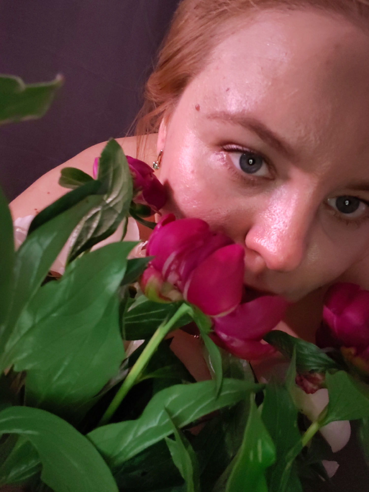 A close-up, top-down shot features a person with fair skin and light brown hair peering through a bouquet of pink peonies and green leaves. The person's right eye is clearly visible, with a blue iris and a small, dark mole on their forehead just above the eyebrow. They are wearing a gold earring with a green stone in their right ear. The background is a dark, solid color. The image was taken in Izhevsk, Russia.