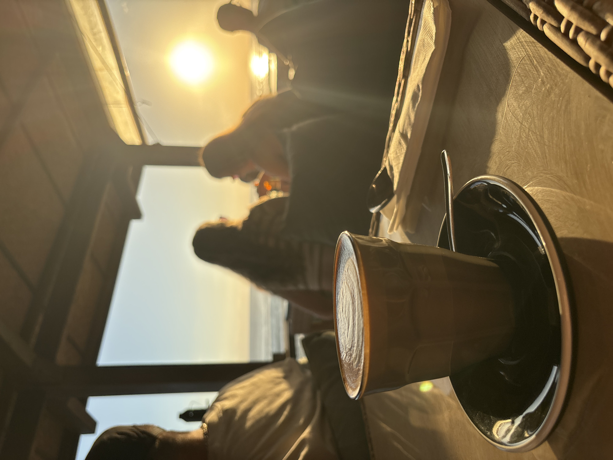 The image shows a close-up of a coffee cup and saucer on a table. A spoon is in the saucer, and the cup is filled with coffee. In the background, there are blurred figures of people sitting at the table, with the ocean visible in the distance through a window. The sun is shining brightly, casting a warm glow over the scene. Based on the prompt the location is North Kuta, Indonesia.