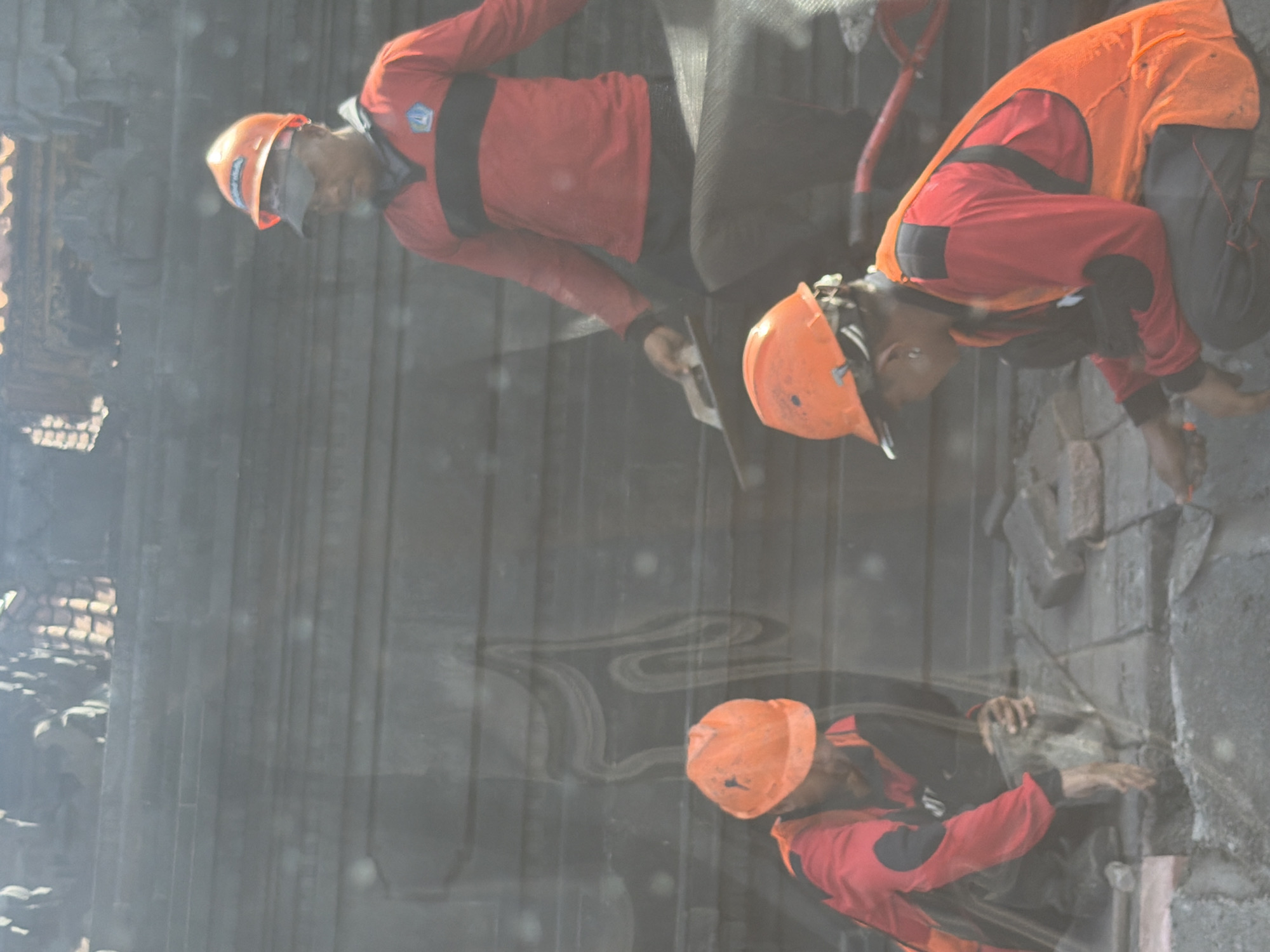 Three workers in hard hats and uniforms are engaged in restoration or maintenance work on a stone structure, likely an ancient temple or building, in an outdoor setting during daytime. The image is taken through a slightly hazy or reflective surface, causing some blurring and reflections.
The workers are wearing orange hard hats and red long-sleeved shirts with black accents. One worker in the foreground, bending over, also wears an orange safety vest. This worker appears to be examining or working on the intricate stone carvings of the wall. Another worker, standing upright with a tool (part of a red shovel handle is visible), is positioned behind the first. A third worker is visible further back, also wearing a red shirt and orange hard hat.
The background consists of dark, textured stone, suggesting substantial masonry work or elaborate architectural features common to temples in the region. The workers appear focused on their tasks. Text visible on one hard hat reads "BAHANA," and a small blue and white logo is present on one worker's red shirt. The scene hints at ongoing efforts to preserve or repair historical or cultural sites.