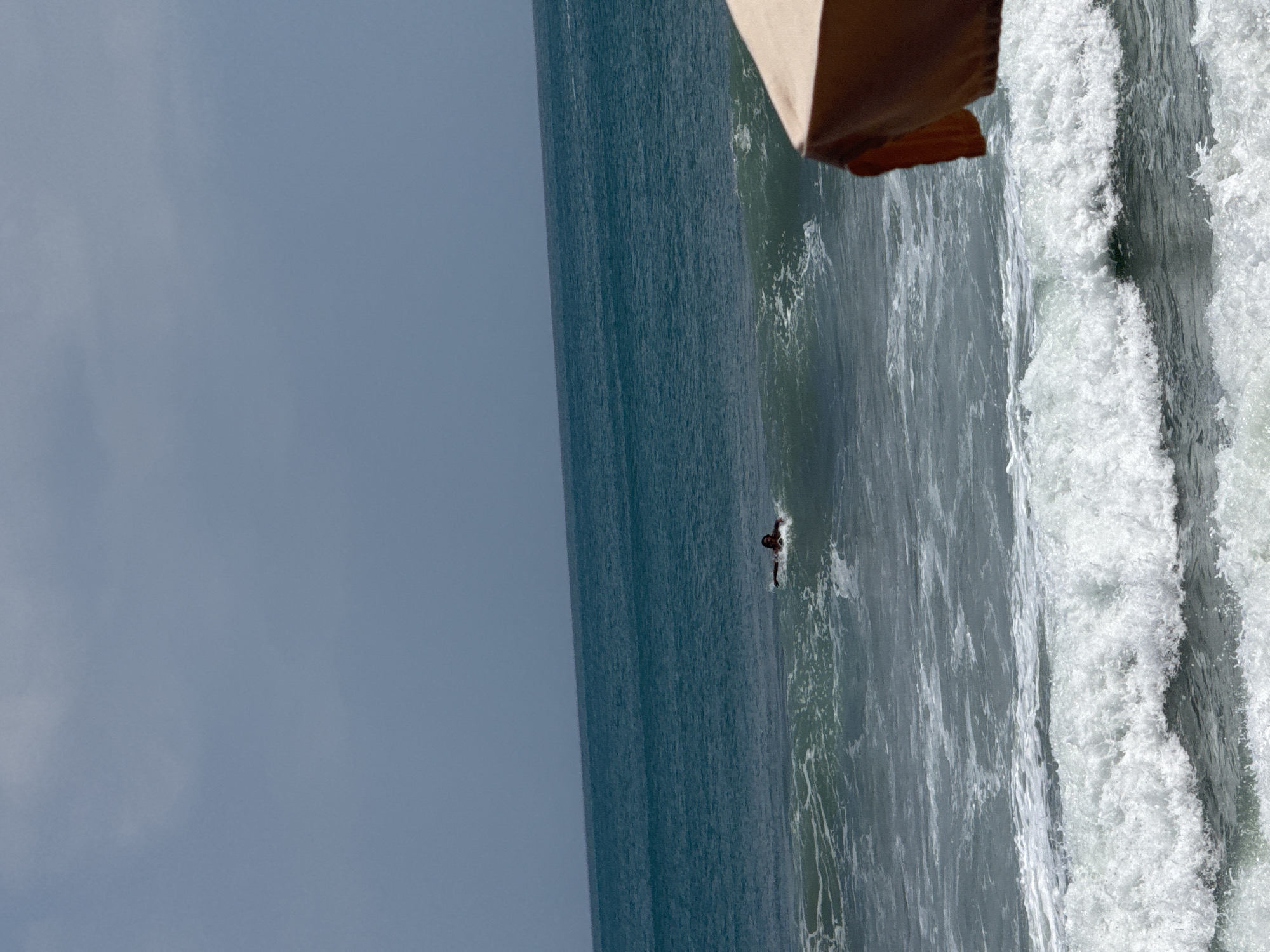 An overhead shot captures the vast expanse of the ocean with a single individual swimming in the middle distance. The water is a deep teal, transitioning to lighter turquoise near the shore where waves are breaking. A light brown beach umbrella is partially visible in the foreground, adding a splash of color to the scene. The sky is a muted gray, suggesting an overcast day. The image appears to be taken on a beach in Kuta, Indonesia, a popular tourist destination known for its surfable waves and vibrant atmosphere.