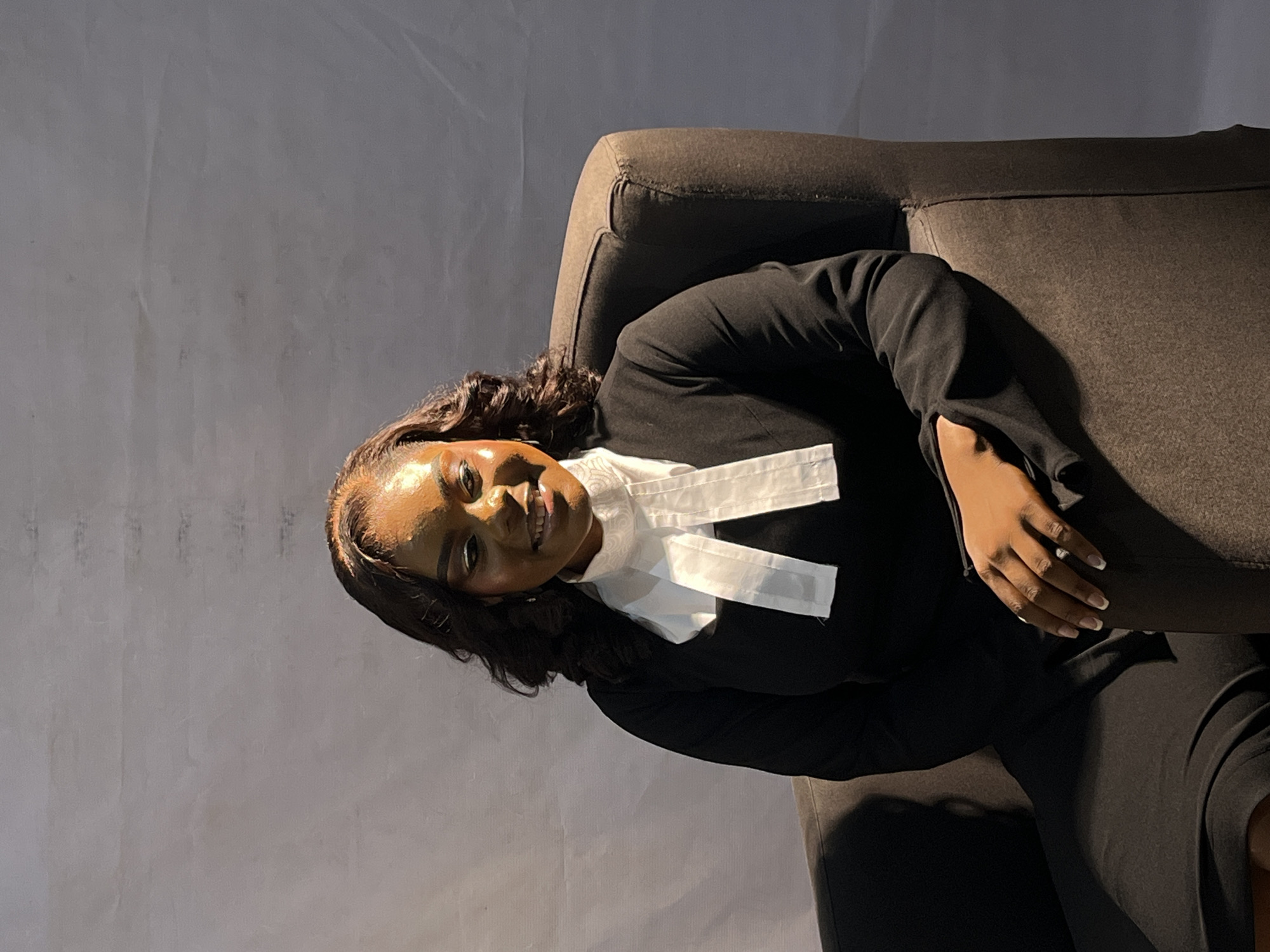 A medium shot captures a Black woman seated in a dark brown armchair against a plain, light gray backdrop. She is looking directly at the camera with a slight smile. She has dark, wavy hair styled to frame her face, and is wearing a black robe with a white jabot around her neck. Her makeup is noticeable, with a bold eyeliner. The lighting casts a warm glow on her face and hair, with some shadows visible on the backdrop. The armchair has a textured fabric. Her left hand rests on her lap, showing manicured nails. The lower part of the image shows the dark fabric of her robe and skirt.