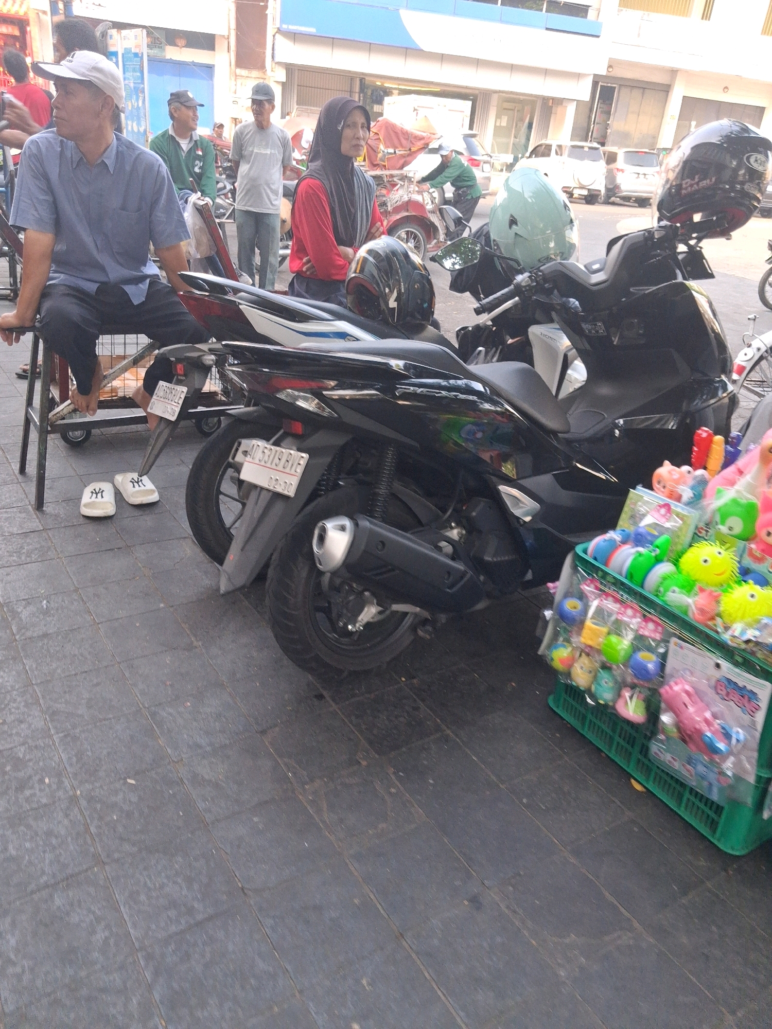 The image depicts an outdoor scene likely in the city of Surakarta, Indonesia, given the Indonesian license plates on the motorcycles. In the foreground, a black motorcycle is parked on a tiled sidewalk. To the right of the motorcycle, a green plastic crate is filled with colorful toys, some of which appear to be squeeze toys and bubble blowers. The crate is positioned on the sidewalk, suggesting a street vendor stall.
In the background, several people are visible. A man wearing a light blue shirt and a white baseball cap is seated on a metal frame structure, facing left and looking away from the camera. Next to him, a woman in a red long-sleeved shirt and a black hijab is standing. Further behind them, two men are standing. One is wearing a grey t-shirt and a dark cap, while the other wears a green long-sleeved shirt and a black cap. Other individuals and elements like the side of a building, a partially covered vehicle, and a red and white structure are also present in the background, indicating a bustling urban environment. The overall scene suggests a street-side commercial area.