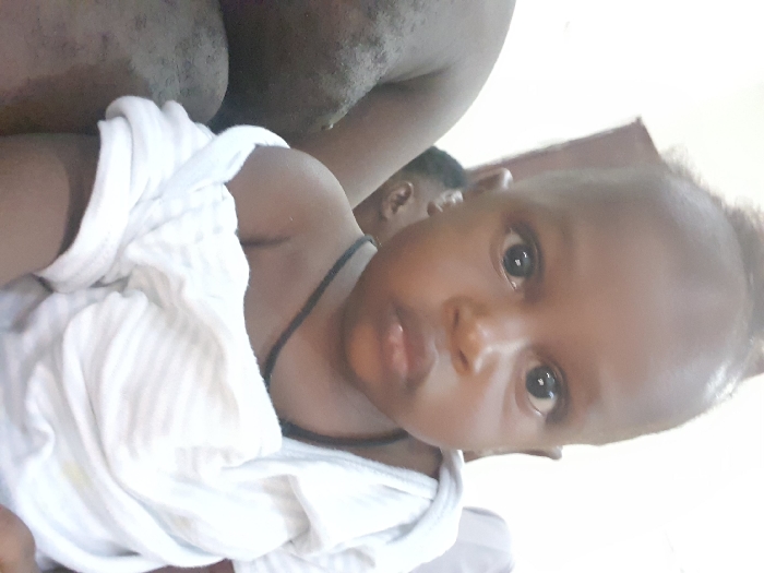 A front-facing close-up portrait depicts a young child, likely an infant or toddler, with dark skin and dark eyes, gazing directly at the camera. The child exhibits short, dark hair, styled with a small prominent tuft. The child wears a white, possibly striped, short-sleeved garment, which is displaced from the right shoulder, exposing the skin. A thin black cord or necklace is visible around the child's neck. To the left of the child, the bare arm and shoulder of another individual, with dark skin, are partially visible. A third individual's head, specifically an ear, is partially visible in the background to the right of the child's head. The immediate background consists of a light-colored wall with a darker vertical architectural element. The scene is indoors. The precise location within the city of Unknown, Unknown is not discernible from the provided image.