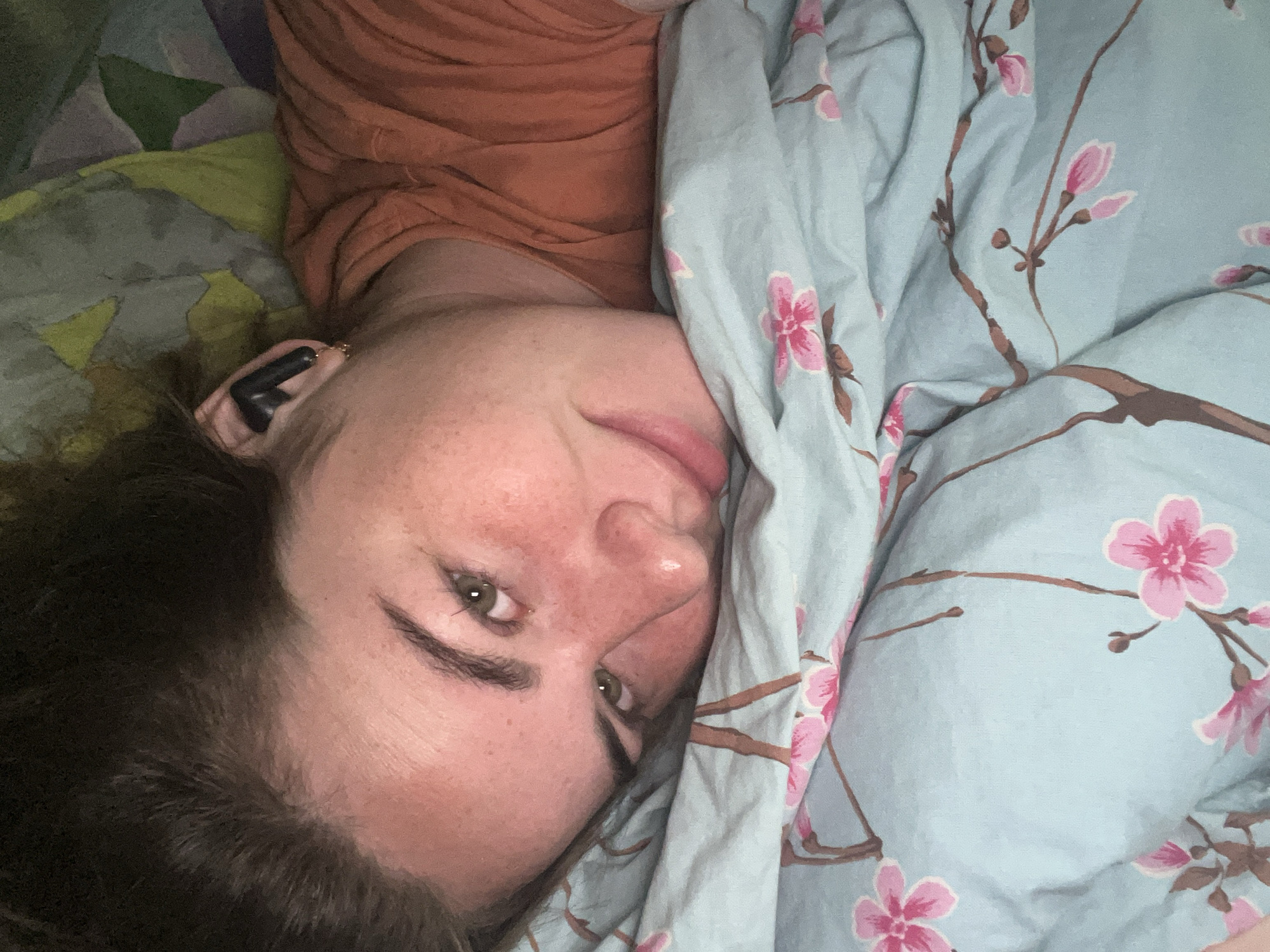 The image shows a close-up of a person lying down on a light blue bedsheet with a floral pattern. The person is looking at the camera with a slight smile. They are wearing an orange t-shirt and have an earbud in their right ear. The lighting is dim, suggesting it might be nighttime or a dimly lit room. The background consists of a pillow with a colorful, abstract pattern and the person's dark hair. The overall setting appears to be a bedroom. There is no identifiable information to determine the specific location in Chaikovsky, Russia.