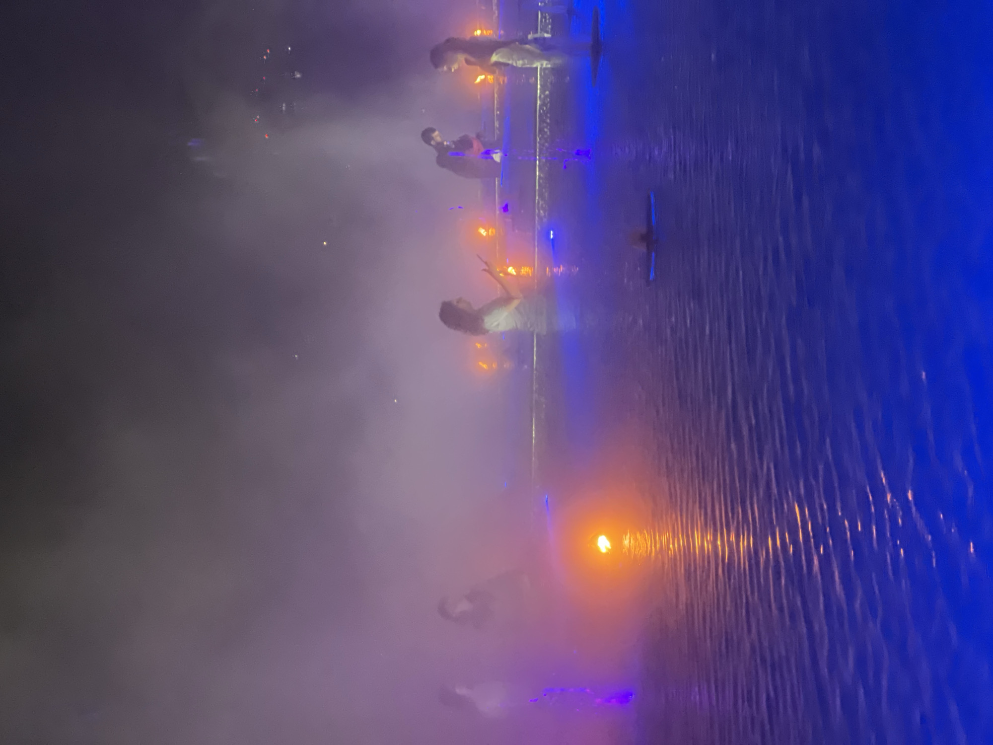 This is a nighttime exterior shot of an event featuring several individuals in what appears to be a shallow body of water illuminated by dramatic purple and blue lighting, with fog or smoke adding to the atmosphere. There are multiple figures, some of whom appear to be dancers or performers, dressed in light-colored, flowing garments. One figure in the center is playing a stringed instrument. Small, orange flames or lights are visible in the background, possibly part of the stage design. The water has ripples and reflections of the lights. This scene is located at the "Parus" (Sail) resort in Krasnodar, Russia.