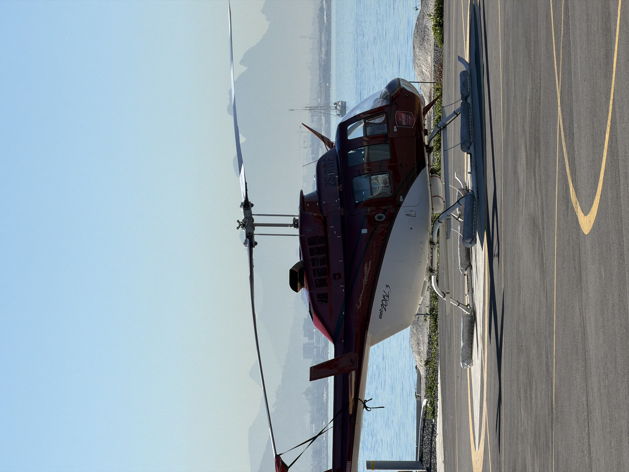 A burgundy and white Bell 206L3 helicopter is parked on a gray asphalt surface with yellow lines, possibly a helipad or apron at a landing strip. The helicopter faces toward a body of water, beyond which are mountains shrouded in haze, suggesting a coastal or lakeside environment. The sky above is a pale blue. The helicopter is detailed, with visible rotor blades and landing gear. A strip of green vegetation separates the asphalt from the water. In the distance, faint outlines of boats or ships are discernible on the water. The setting appears to be daytime with good visibility. While the exact location cannot be determined, the scene is not in Cape Town.