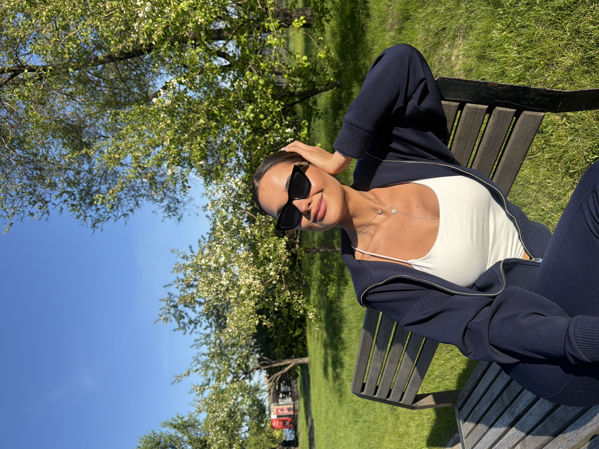 A woman wearing sunglasses, a white tank top, and a dark blue zip-up jacket is sitting on a wooden park bench on a sunny day. She has her hand on her head and is smiling. Trees with white flowers are visible in the background. The location is Krasnoyarsk, Russia.