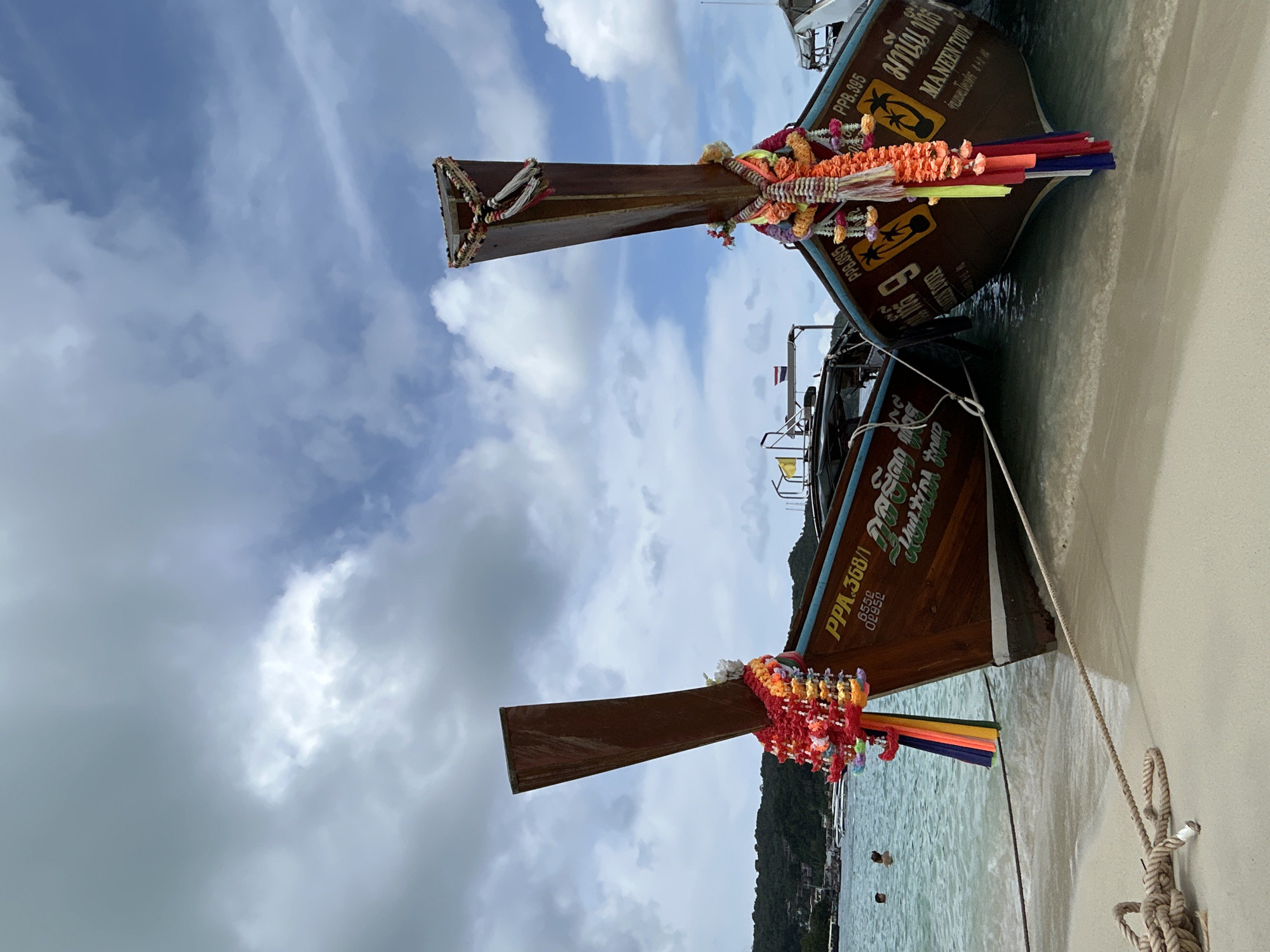 Two brown longtail boats are docked on a sandy beach in Ao Nang, Thailand. The boats are adorned with colorful garlands of flowers and tassels, traditional decorations for Thai boats. The boat on the left has the registration number "PPA.368/1" and some Thai writing. The boat on the right has "PPB.395" and "MANEEN TOUR" written on it, along with Thai text. The water is a clear turquoise color, and the sky above is partly cloudy with a mix of blue sky and white clouds. In the distance, a green, hilly coastline with buildings is visible. Two people are in the water near the shore. A thick rope is coiled on the sand in the foreground, connecting to one of the boats.