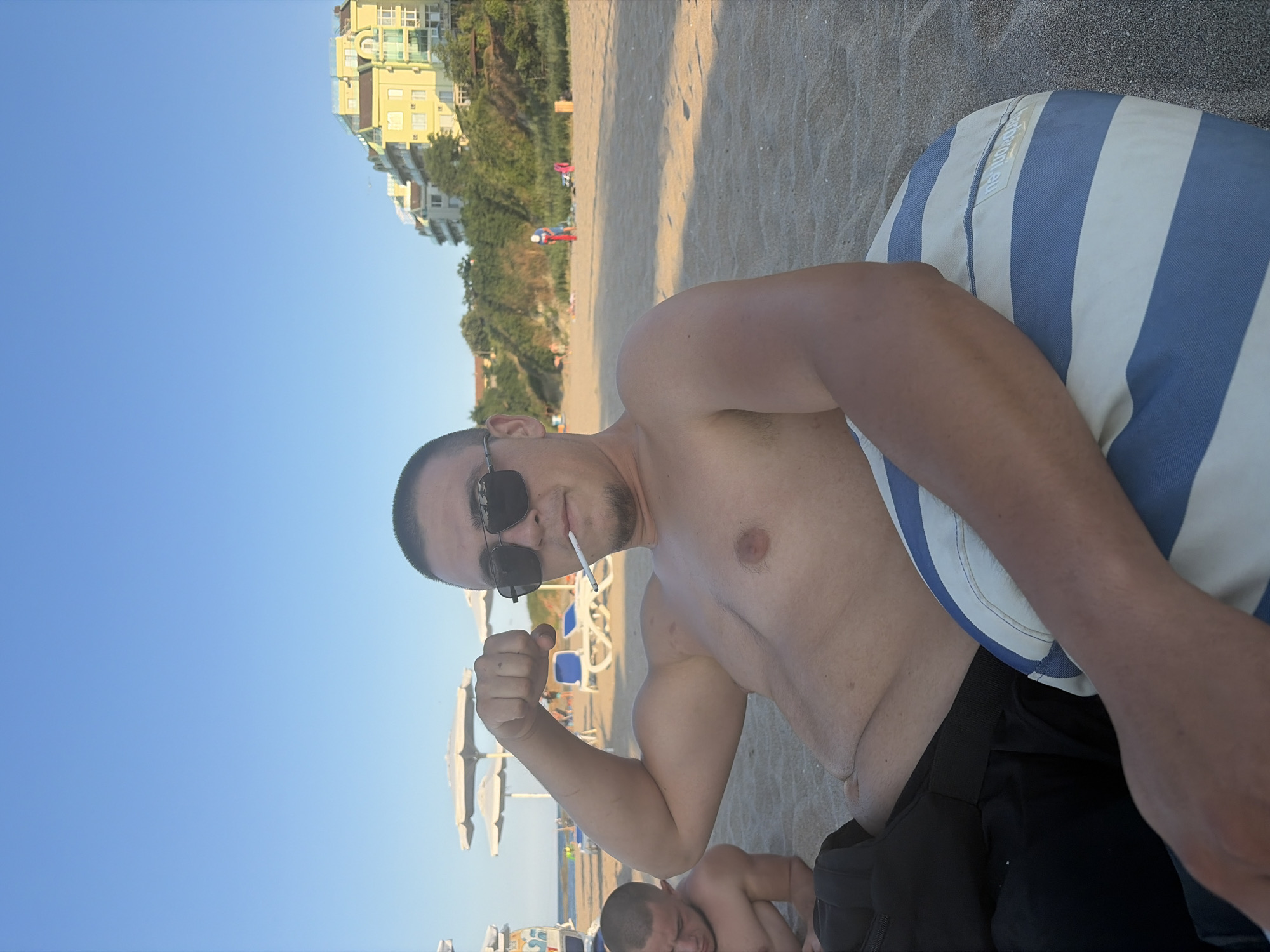 A shirtless man with short hair and sunglasses is sitting on a striped cushion on a sandy beach, with a cigarette in his mouth. He is flexing his bicep. Another shirtless man is visible in the background to the left. In the distance, there are beach umbrellas, lounge chairs, and a multi-story yellow building on a hill overlooking the beach. The sky is clear and blue. The location is the beach in Lozenets, Bulgaria.