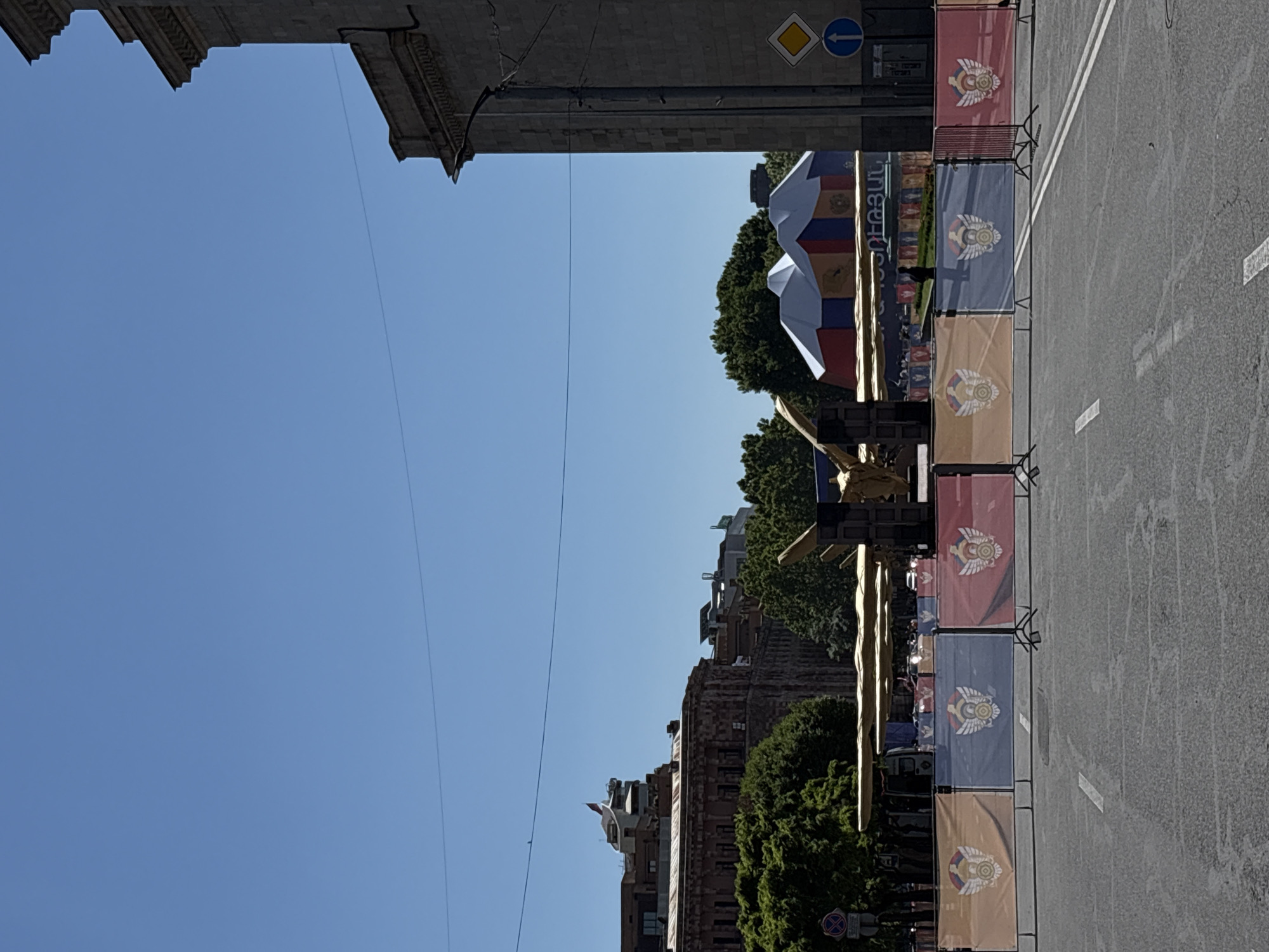 The image shows a street scene in Yerevan, Armenia. A street is lined with decorative banners featuring a golden emblem, likely for an event or celebration. A monument or large decorative installation, potentially related to Armenian culture or history, is visible. Buildings are on either side of the street. A blue sky is overhead.