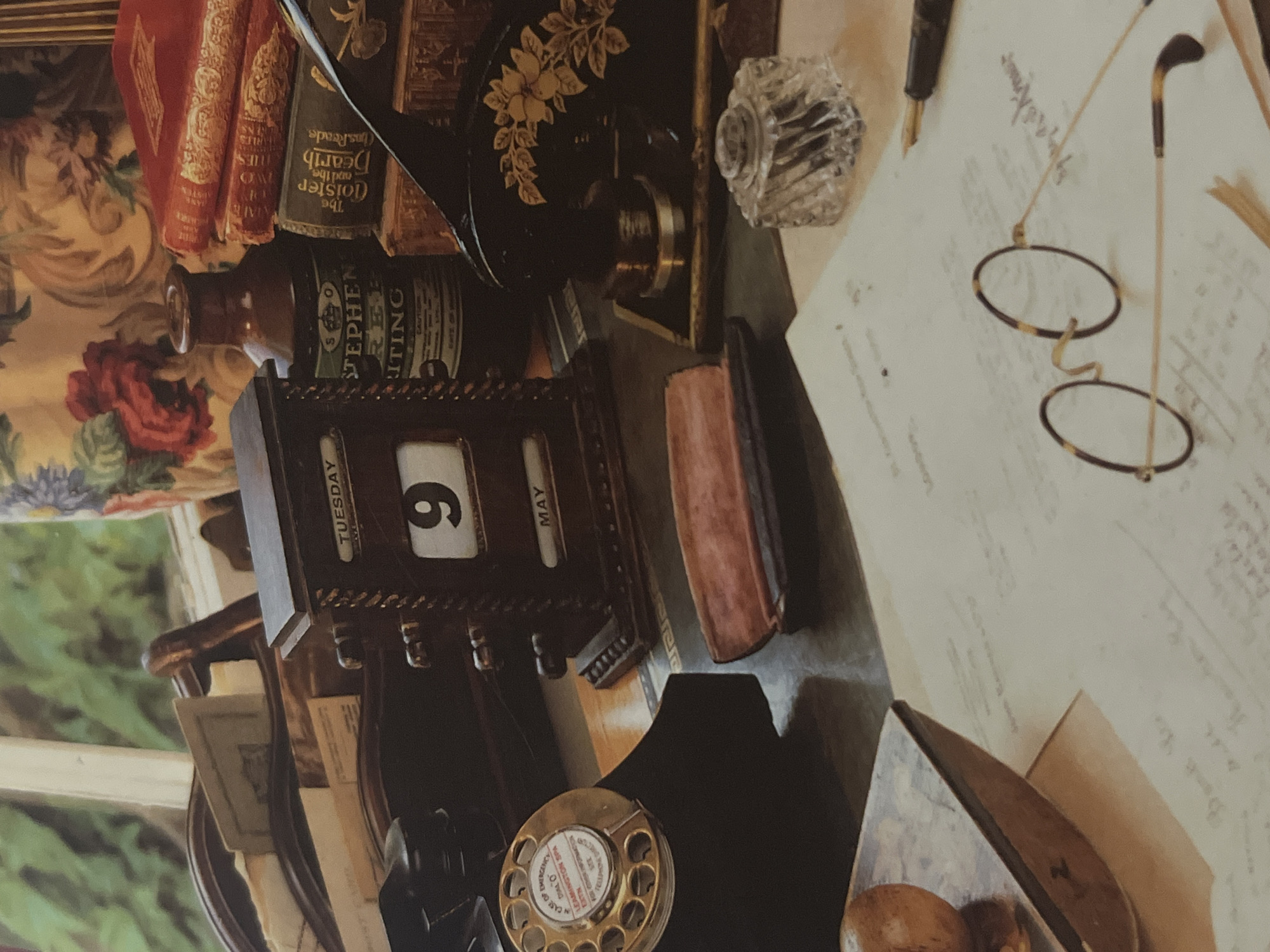 This media file captures a close-up, still life arrangement on a desk, evoking a sense of historical study or writing. The scene is densely populated with antique objects. Prominently featured are several leather-bound books, one with the visible text "The Cloister and the Hearth" and author "Charles Reade." A dark, ornate object, possibly a sewing machine or decorative embellishment, sits behind a glass inkwell, adding to the antique aesthetic.
In the foreground, a vintage calendar display shows "TUESDAY" and the number "6" with "MAY" also visible. A dark wooden frame surrounds this calendar. To the right, a pair of tortoise-shell framed eyeglasses rests on a sheet of paper filled with handwritten script. A fountain pen lies horizontally nearby. Another object, a dark, upright phone receiver, is partially visible at the bottom left, next to a round, metallic object with holes, resembling the dial of an old telephone, which displays text like "EMERGENCY DIAL O" and "LEAMINGTON SPA." A brown glass bottle with a label that appears to read "STEPHEN WRITING" is also present.
The background includes a richly patterned fabric with floral motifs and a hint of green foliage, suggesting a window or a decorative backdrop. The lighting is somewhat diffused, creating a warm and intimate atmosphere, though the specific time of day or weather conditions are not discernible. The overall impression is one of a meticulously curated collection of items related to writing, reading, and perhaps a past era of communication.
