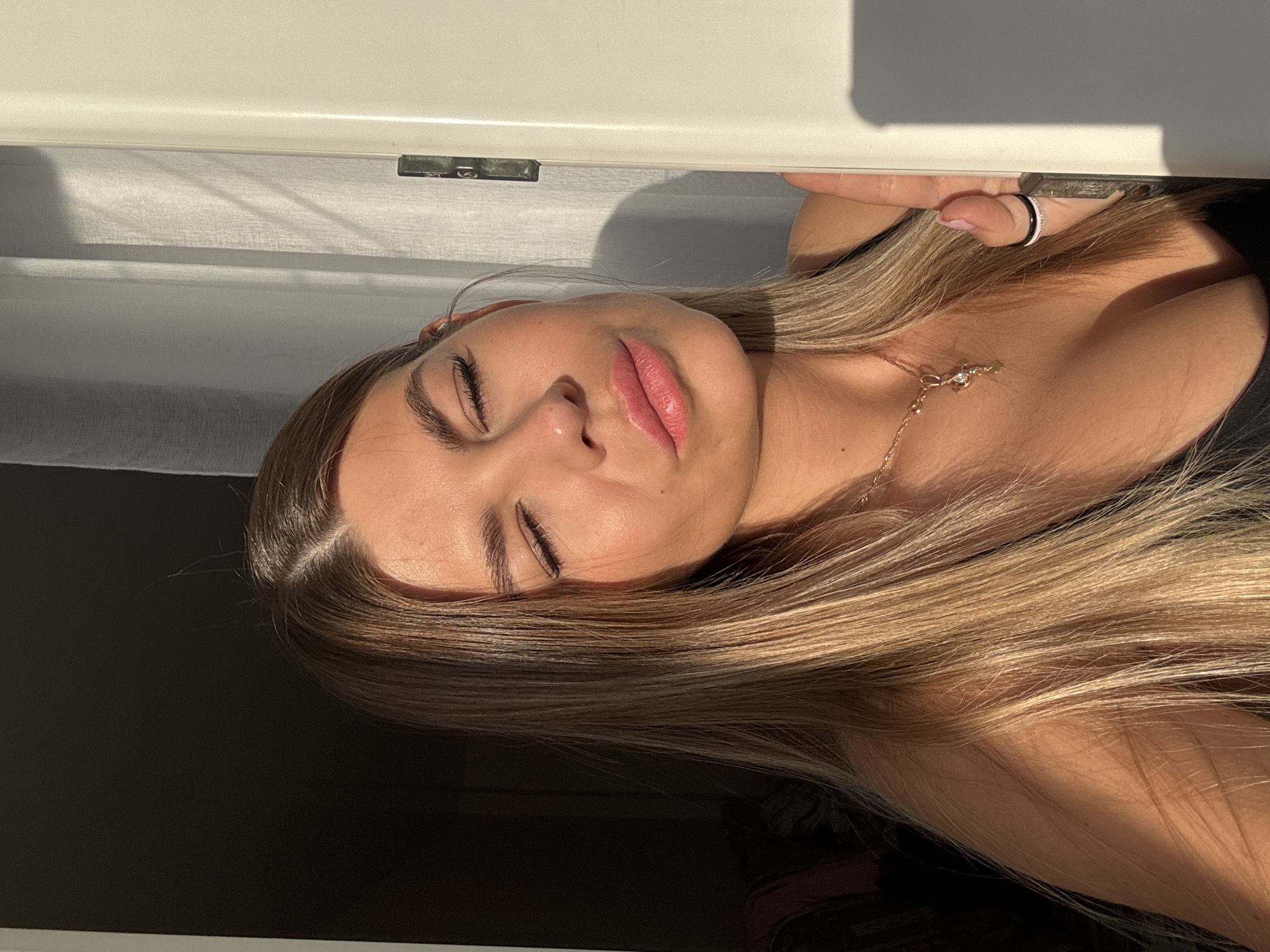 The image shows a close-up of a young woman with long, blonde hair. She is wearing a black top and a necklace with a cross pendant. The woman's eyes are closed, and she appears to be basking in sunlight, which casts shadows on her face and neck. She is positioned near a doorway or window frame, with soft drapery visible in the background. The lighting suggests it is either early morning or late afternoon. There is no discernible location in the city of Sarapul, Russia, visible in this image.