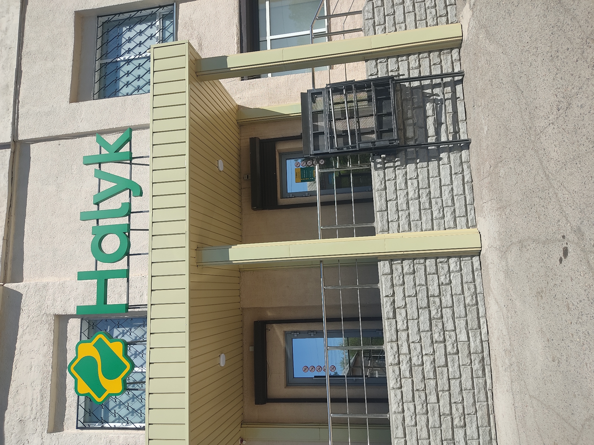 The image shows a building in an urban environment, likely in the city of Unknown, Unknown. The building has a sign that reads "Halyk" in green letters, with a yellow and green logo below it. The building has multiple windows with metal bars on them, and some windows have signs with symbols on them. The building's facade is made of different materials, including a textured concrete, wood panels, and brick-like material. There are metal railings and fixtures attached to the building. The environment appears to be a bright day, with clear skies reflected in the windows. The building is adjacent to a concrete sidewalk.