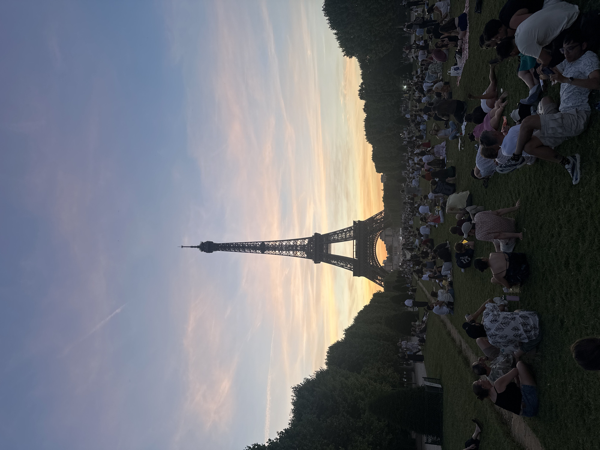 This is a wide-angle, eye-level shot of the Eiffel Tower in Paris, France, during what appears to be sunset. The sky displays a gradient of soft blues, pinks, and oranges, with a single contrail visible in the upper left. The Eiffel Tower, a prominent iron lattice tower, is centered in the frame and dominates the background. In the foreground and mid-ground, a large grassy area is filled with numerous people sitting and lying down, some in small groups, appearing to picnic or socialize. Trees line the sides of the frame, with the one on the left being more prominent and closer to the camera. The lighting suggests late afternoon or early evening, with the sun low on the horizon, casting long shadows and creating a warm, diffused light.