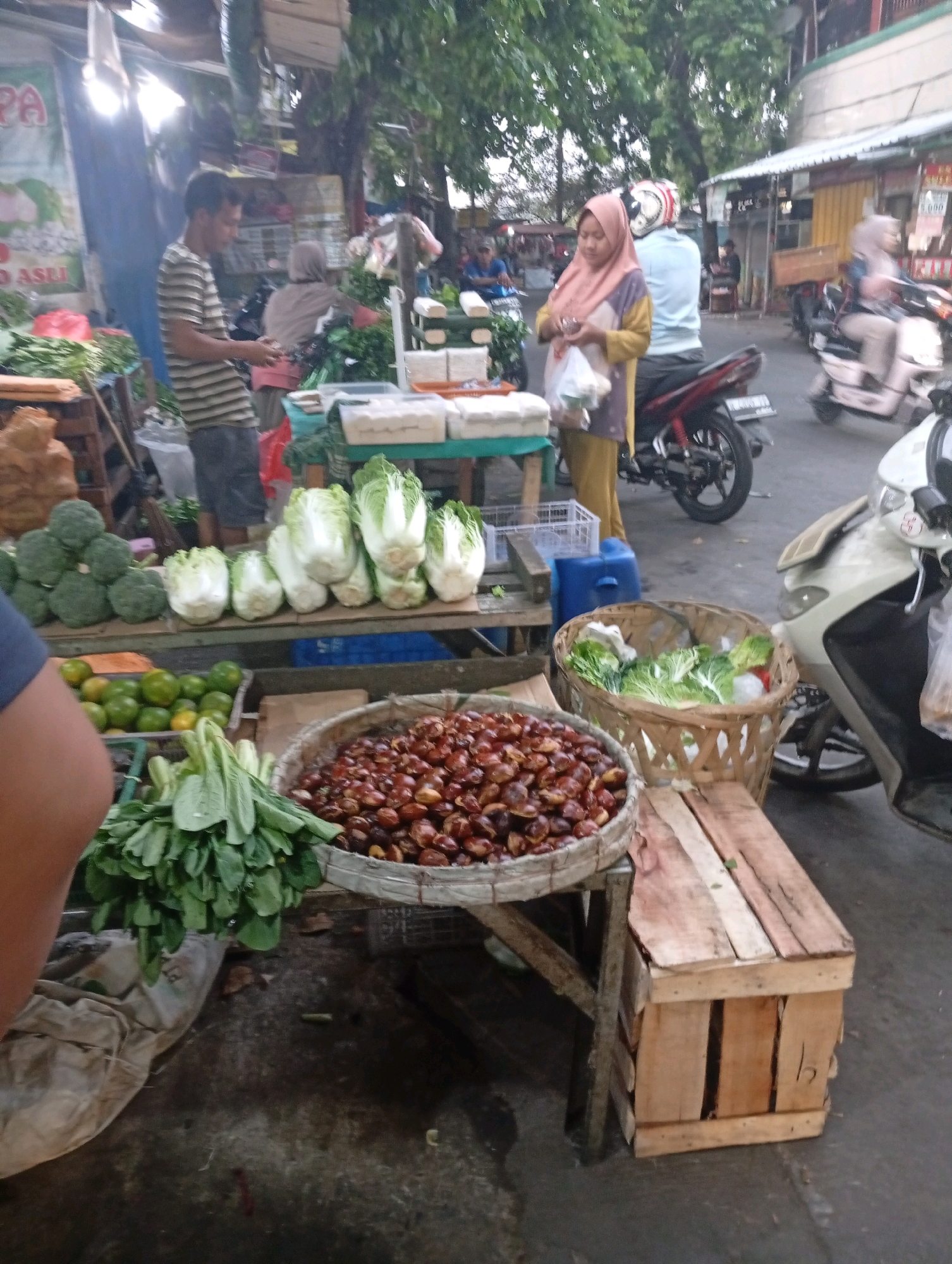 The image depicts a bustling outdoor market scene, likely in North Jakarta, Indonesia. In the foreground, a man in a striped t-shirt and grey pants stands behind a stall laden with fresh produce. To his left, a pile of broccoli is visible, followed by rows of napa cabbage. Further to the right, a basket overflows with green leafy vegetables, and next to it, a large woven basket holds a mound of reddish-brown nuts. A wooden crate sits on the ground beneath the stall.
In the mid-ground, a woman wearing a pink hijab and a yellow and purple outfit is holding a plastic bag. She is standing near a motorcycle. Another person, wearing a light blue shirt and a helmet, is also near a motorcycle. In the background, more market stalls and buildings can be seen, with trees providing shade. The overall atmosphere suggests a typical day at a local Indonesian market.