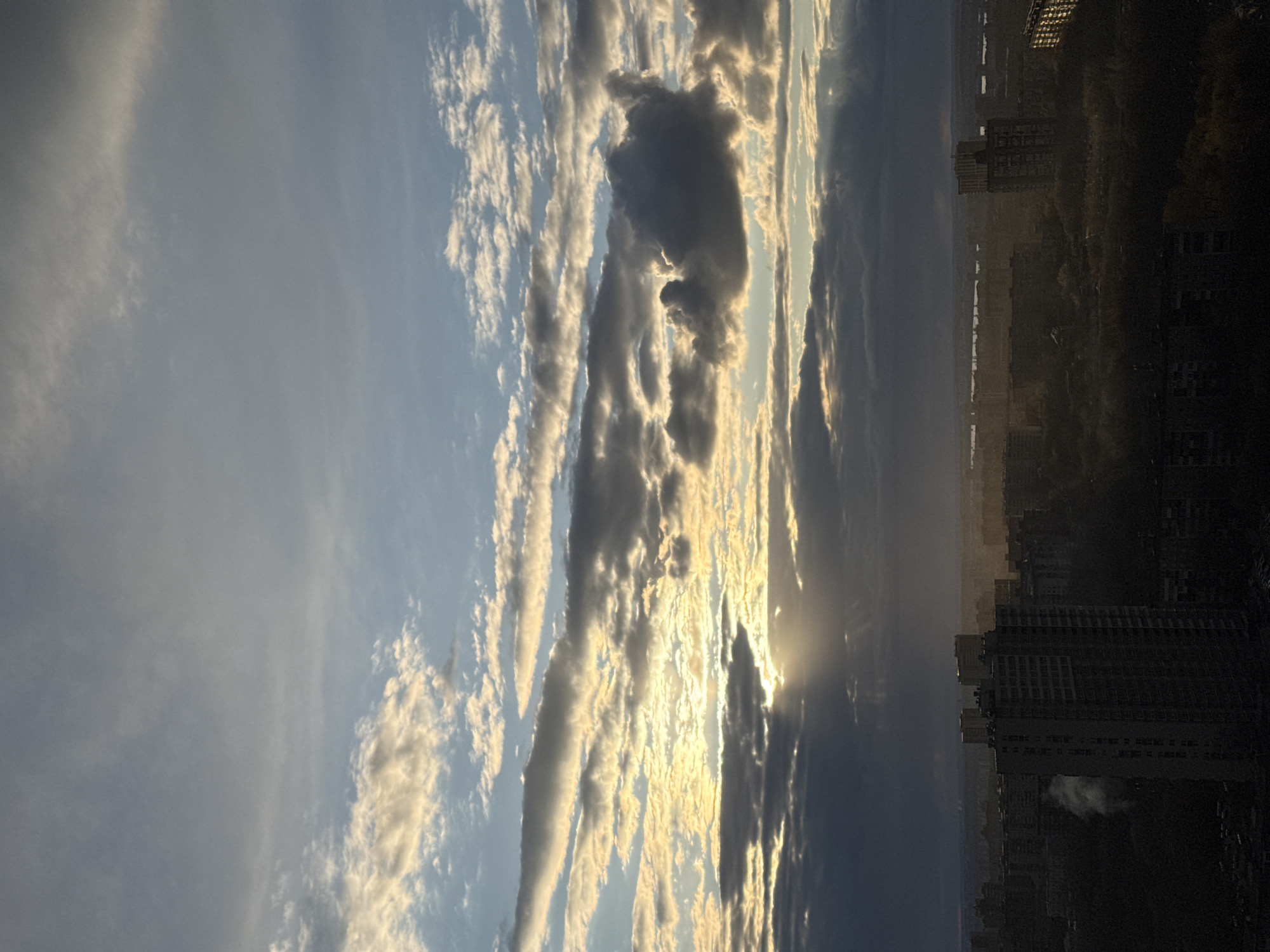 A panoramic landscape photograph captures a dramatic cityscape scene during either sunrise or sunset, viewed from an elevated perspective. The upper two-thirds of the image are dominated by a dynamic sky filled with varied cloud formations. On the left, softer, wispy clouds blend into a light blue sky. Towards the center and right, more substantial, elongated clouds are present, some illuminated with a vibrant golden light from the sun, while others are cast into dark, striking silhouettes.
The sun, positioned low on the right side of the frame, is partially obscured by dense clouds, creating powerful sunrays (crepuscular rays) that pierce through the cloud cover. This intense backlight paints the edges of the clouds with a warm, golden glow, creating strong contrasts between light and shadow across the sky.
The lower third of the image reveals a sprawling city skyline, rendered largely in silhouette. Numerous buildings, ranging from mid-rise to high-rise structures, appear as dark, geometric forms stretching across the horizon. A particularly prominent tall building stands out as a dark, angular shape towards the bottom right. The cityscape extends into the distance, fading into a hazy horizon line that separates the urban landscape from the sky.
The golden light and strong contrasts between illuminated and shadowed clouds and buildings strongly suggest the time of day is either early morning (sunrise) or late afternoon (sunset). The scene conveys a sense of grandeur and tranquility, highlighting the natural beauty of the sky over an urban environment. The location is an urban setting in Perm, Russia, but no specific landmarks or text are visible within the frame, and no human activity or individuals are depicted.