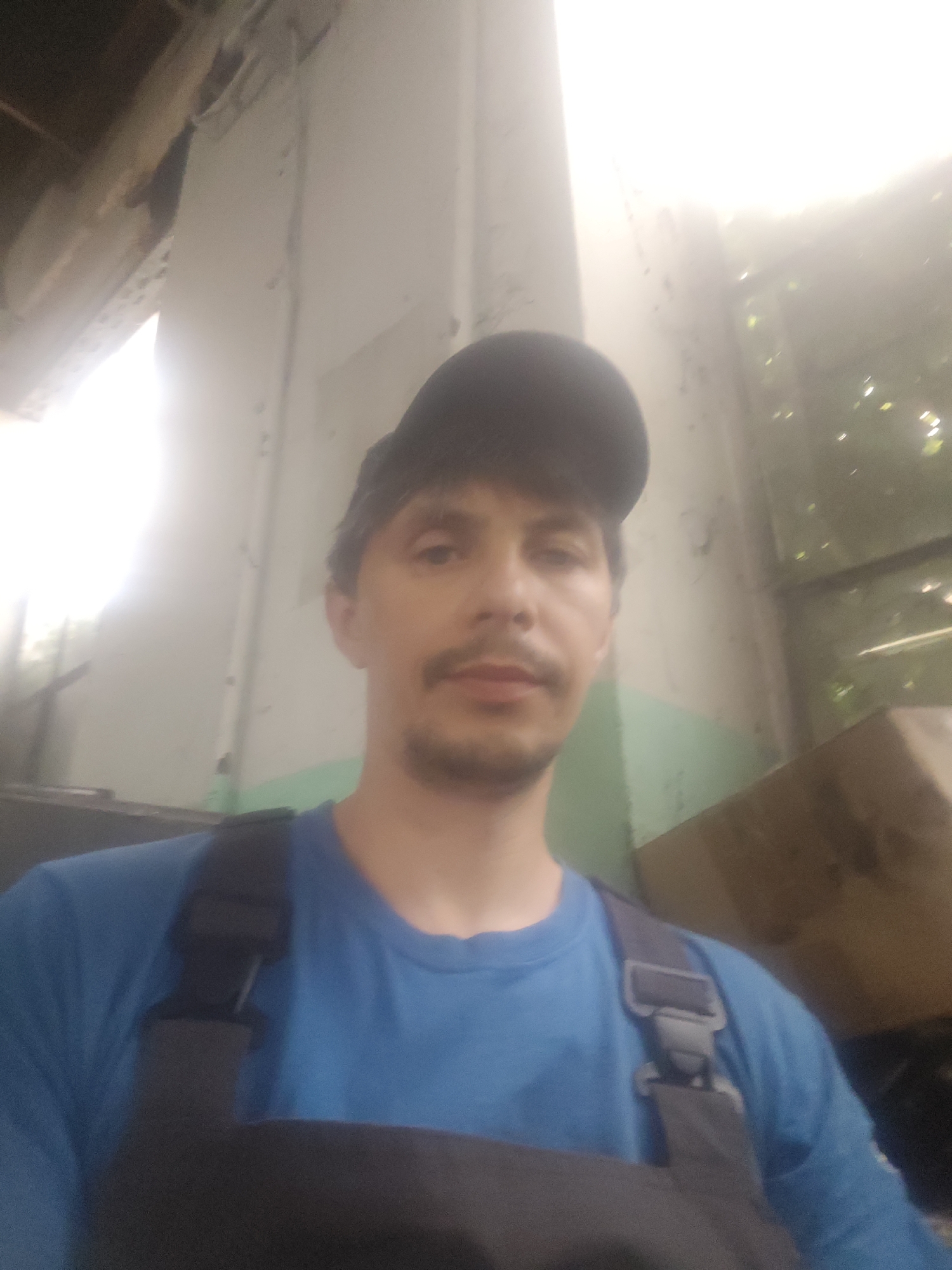 A medium shot selfie captures a man wearing a blue t-shirt and dark overalls, with a dark baseball cap on his head. He has a mustache and a slight goatee. The background appears to be an indoor industrial or dilapidated setting, with exposed beams and a brightly lit area to the left. To the right, a window shows some greenery outside. The overall lighting is bright, with some areas overexposed. The image was taken in Goczałków, Poland.