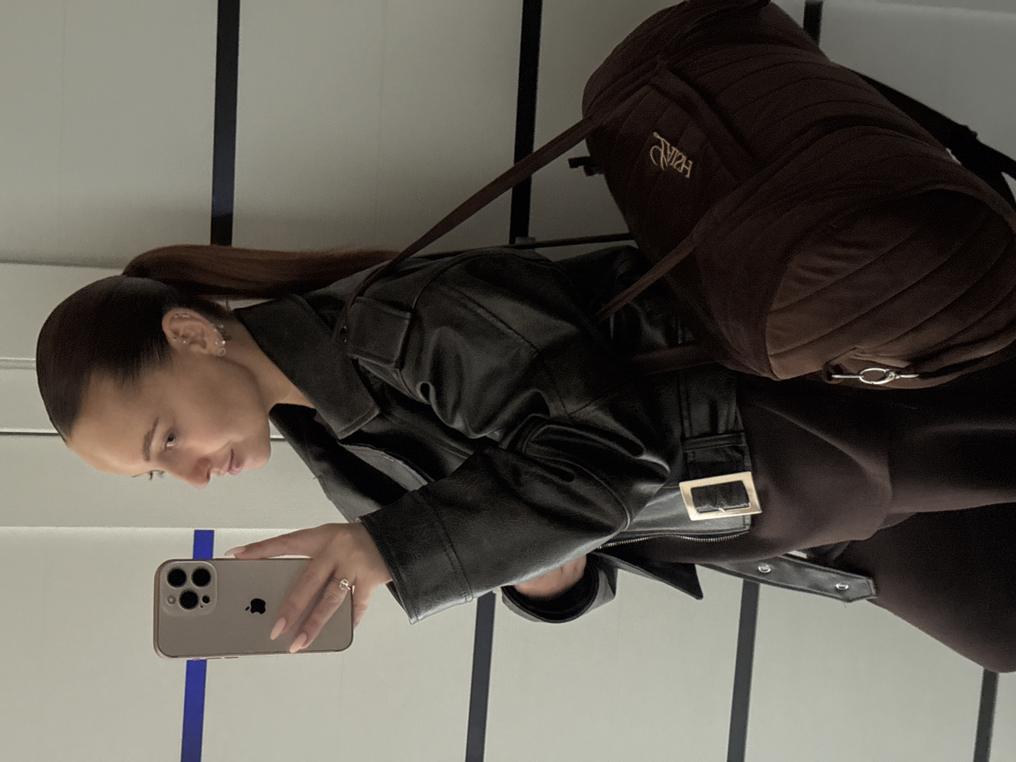 A woman stands in profile, taking a mirror selfie with her phone. She has her hair pulled back in a ponytail and is wearing a black leather jacket and a brown outfit. She is also carrying a brown quilted bag. The background appears to be a modern interior with white walls and horizontal blue and black lines. This scene is located in Chaikovsky, Russia.